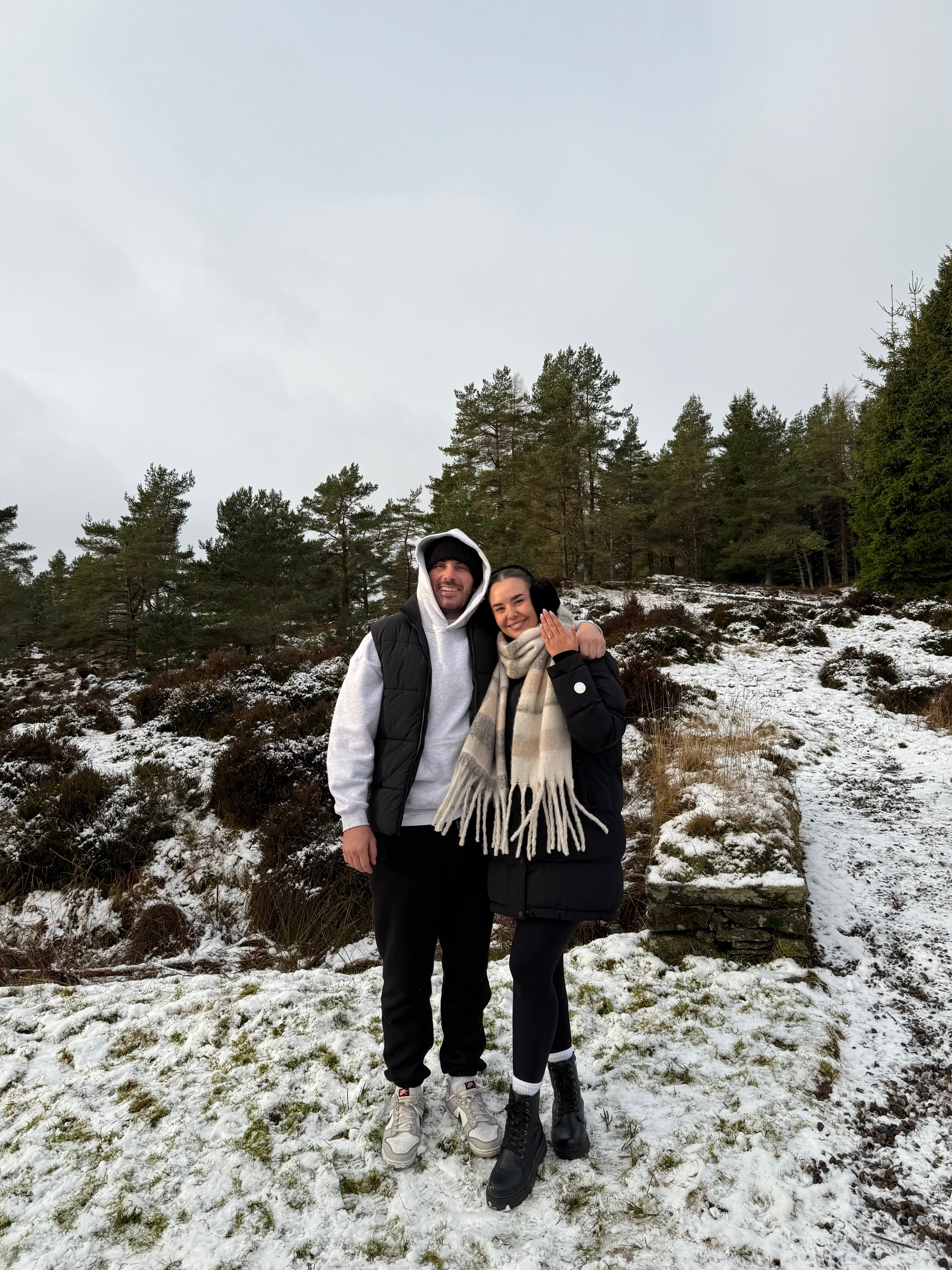 Two people in winter clothing stand on a snowy landscape, with pine trees in the background. The woman wears a scarf and shows a ring.