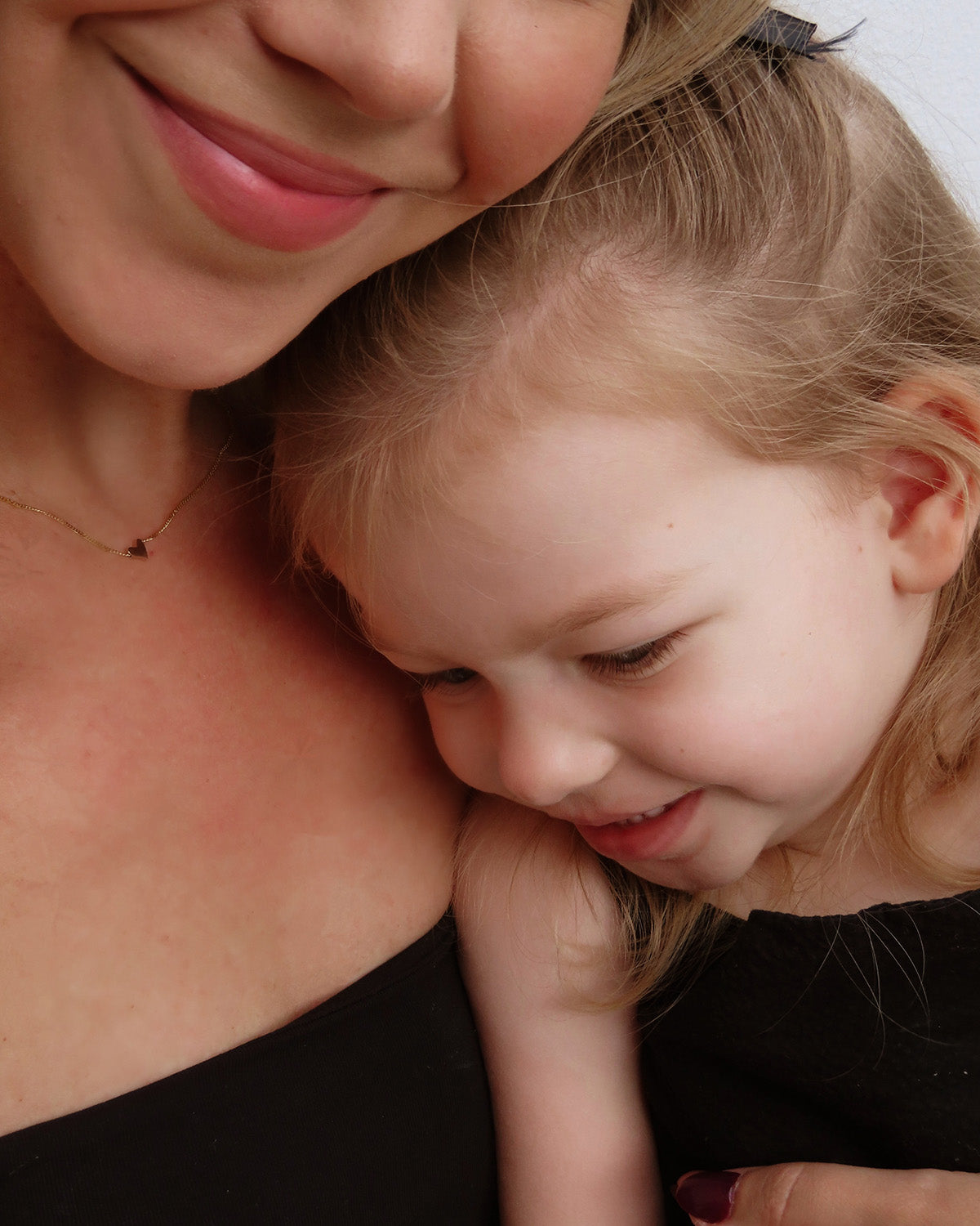 Mother and child wearing black outfits, embracing closely. The mother wears a delicate gold necklace, smiling affectionately.
