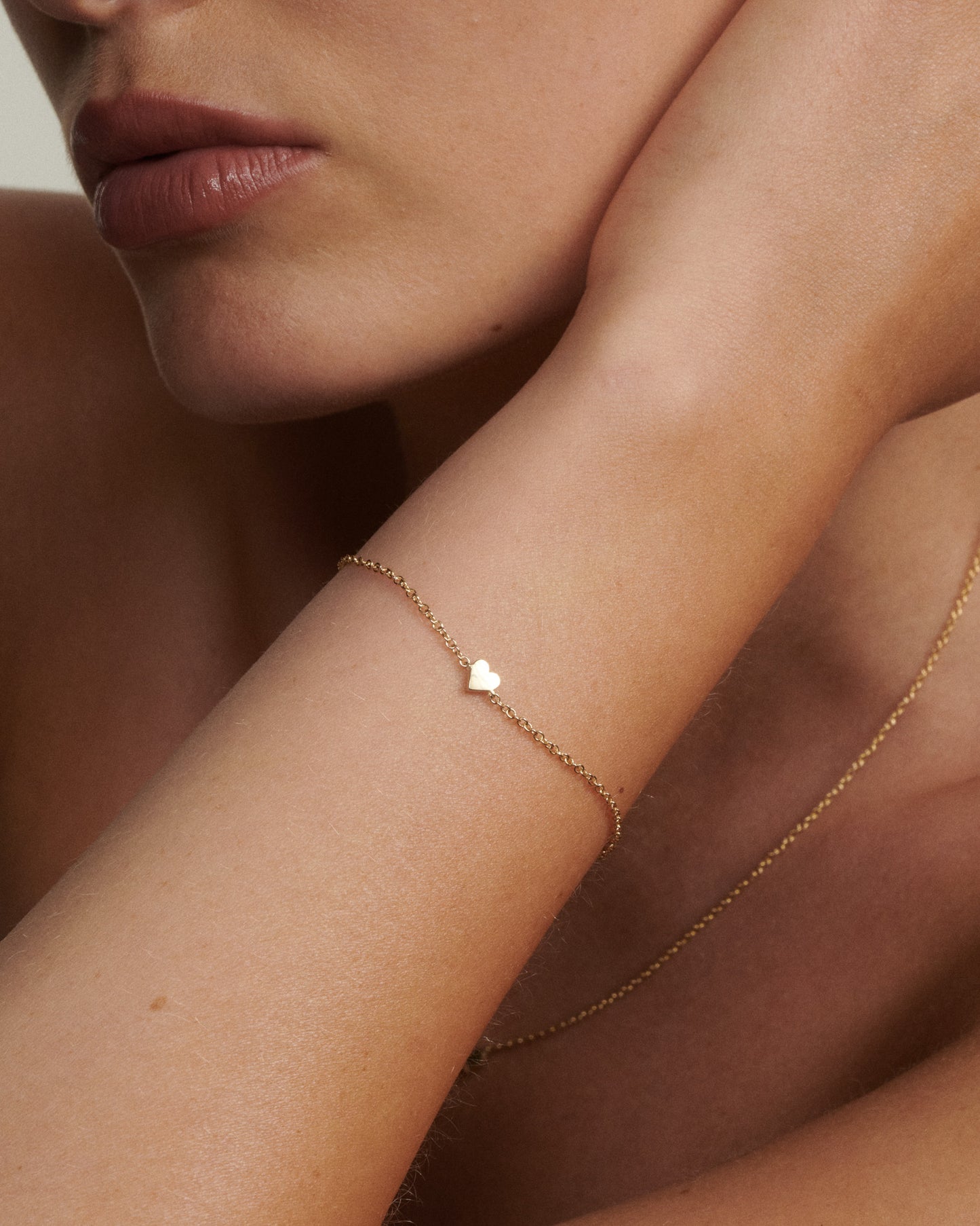 Close-up of a person wearing a delicate gold bracelet with a heart charm on a neutral background