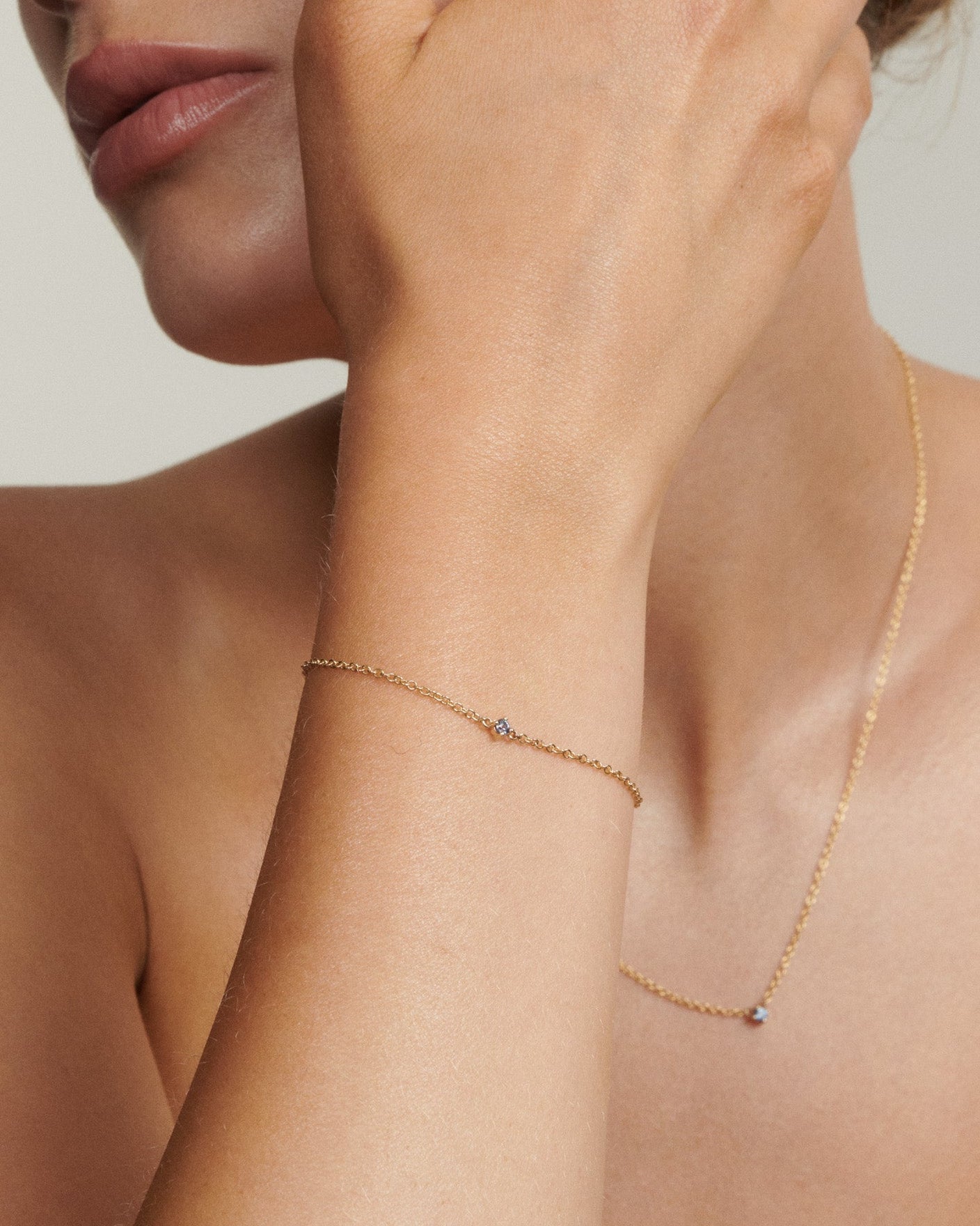 Close-up of a person wearing a delicate gold bracelet and necklace on a neutral background
