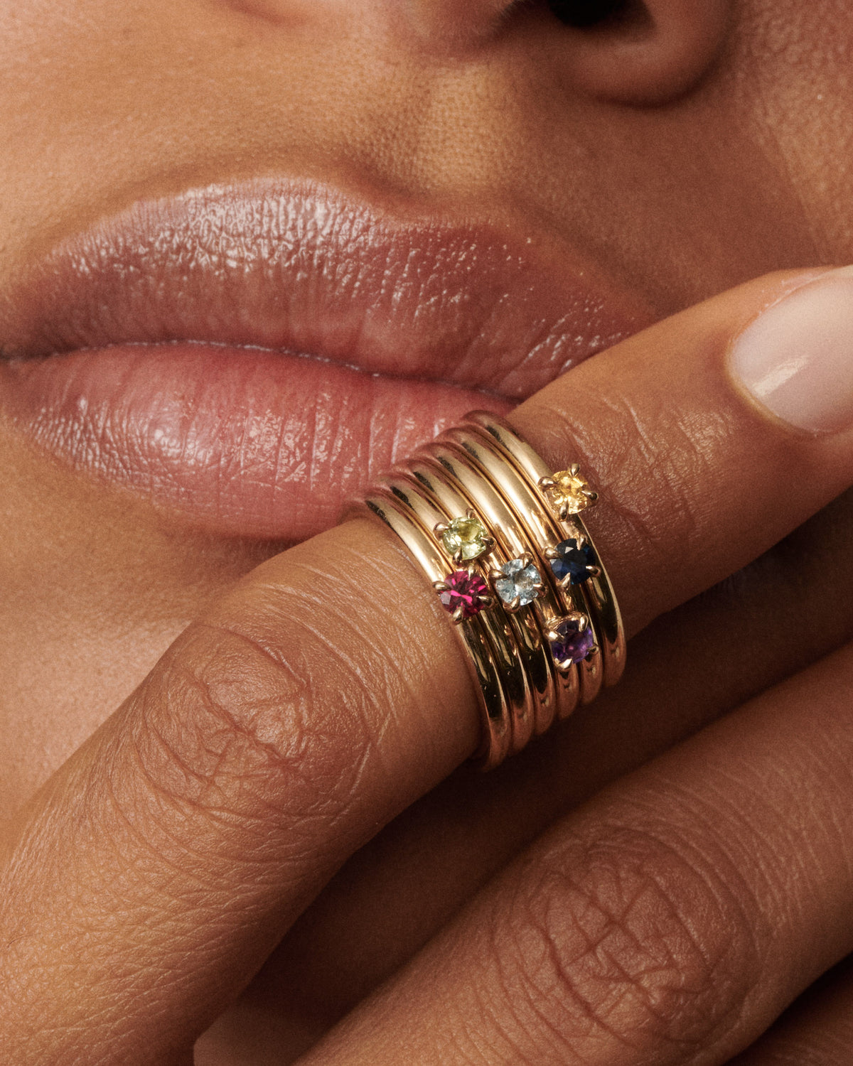 Woman wears a stack of 6 birthstone rings on her finger that is placing on her lips [text overlay: Citrine, Sapphire, Amethyst, Aquamarine, Peridot and Ruby Birthstone Rings (top to bottom)]