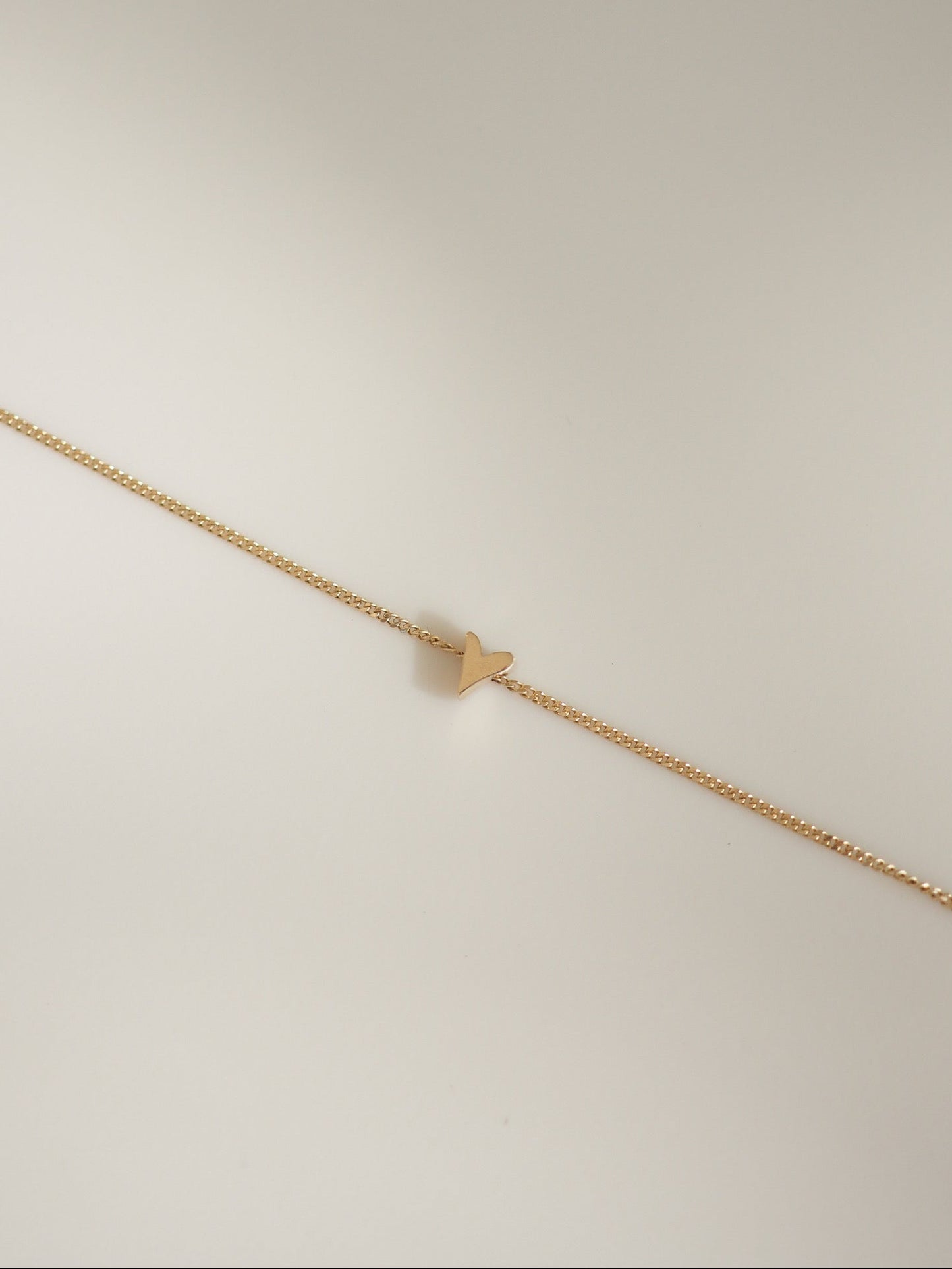 A delicate gold chain bracelet with a small, minimalist gold heart charm, displayed diagonally on a clean, light beige background.