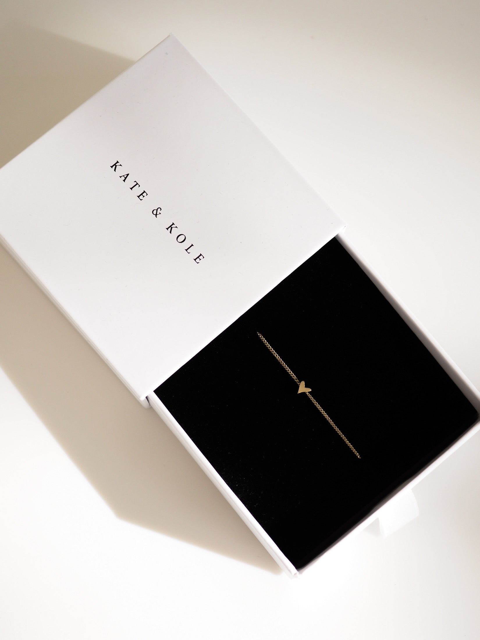 A delicate gold heart-shaped bracelet displayed in an open white presentation box with a black interior. The brand name 'KATE & KOLE' is printed in black on the white lid.