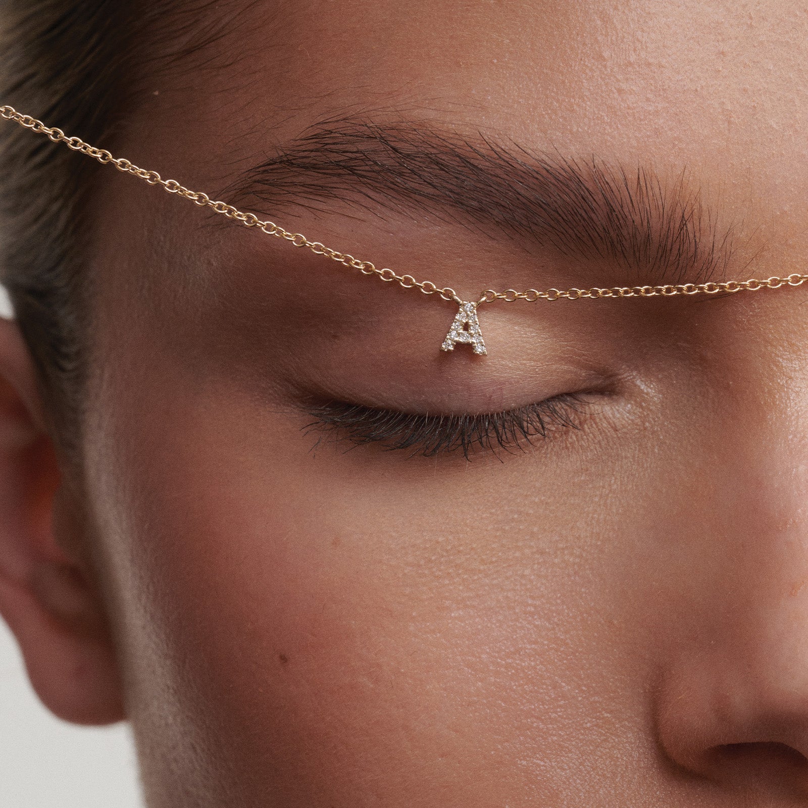 Close-up of a gold chain with a letter 'A' pendant worn across the eye in a creative way.