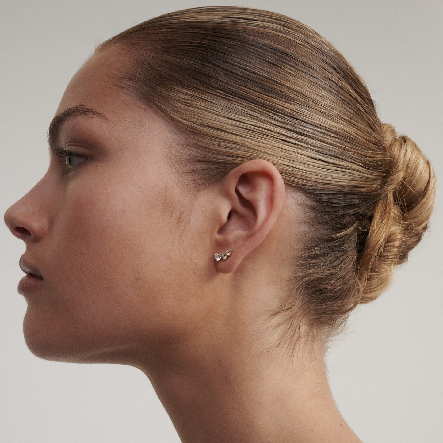 Side profile of a woman with a bun wearing earrings on a neutral background