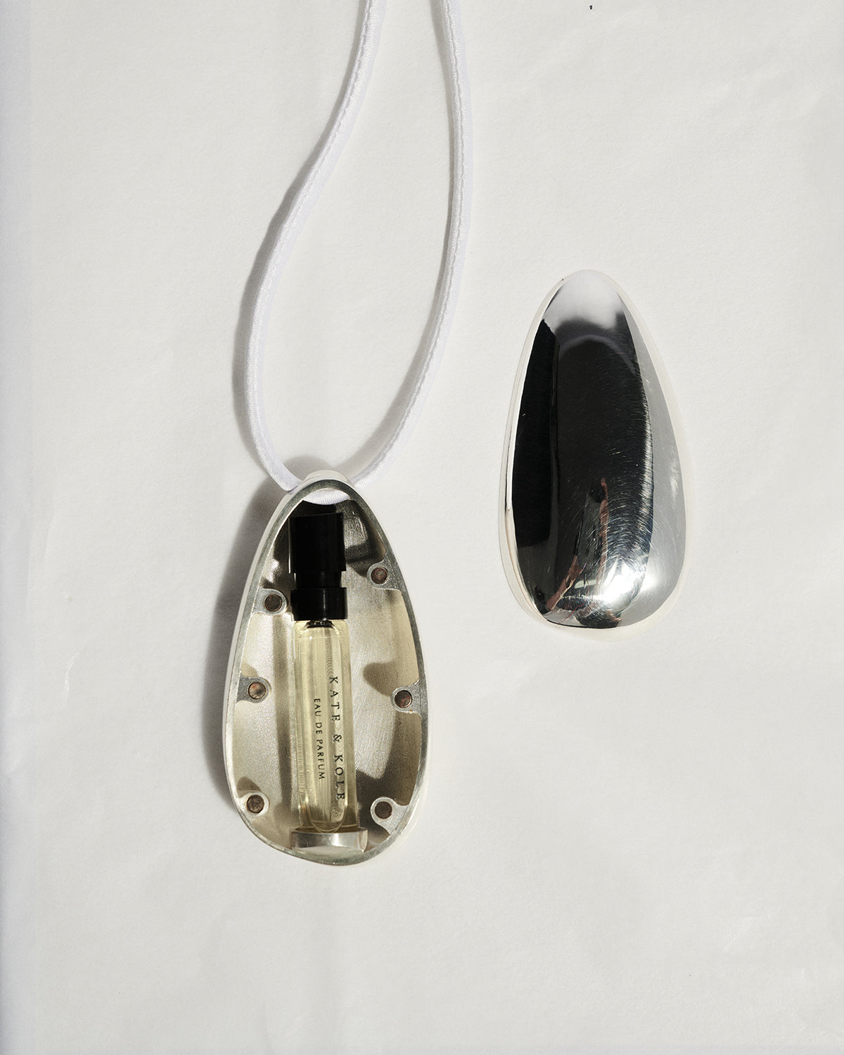 A teardrop-shaped silver pendant necklace shown open on a white background, revealing a small perfume vial inside, with the matching silver lid placed beside it.