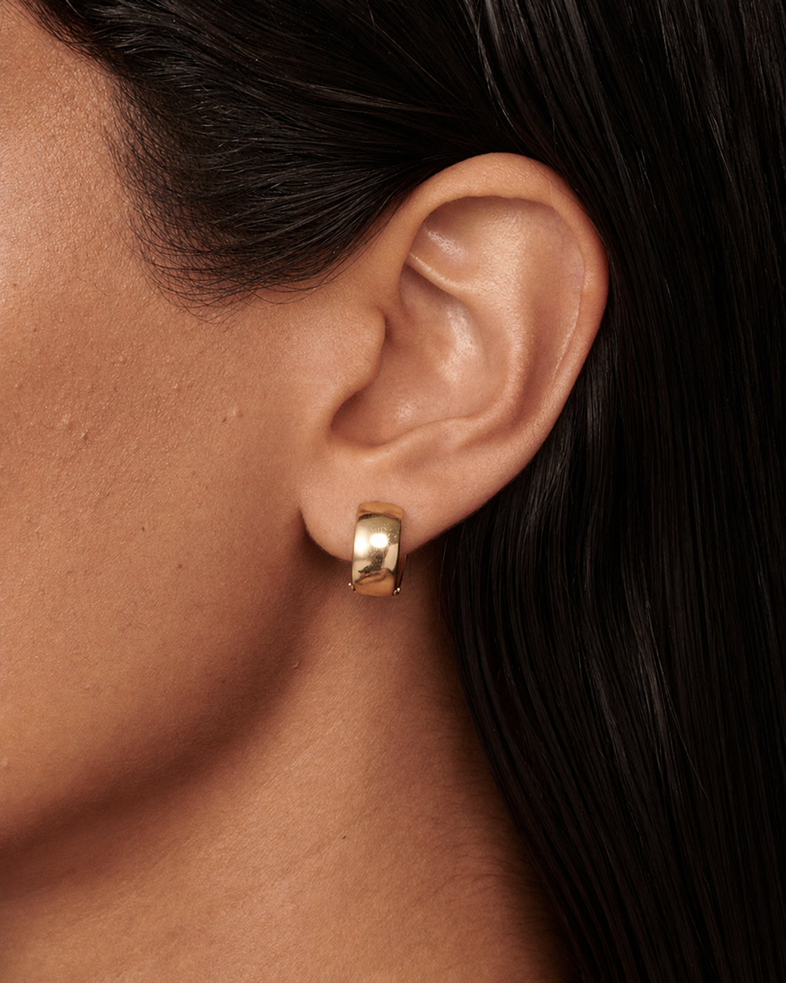 A close-up of a person's ear wearing a small polished gold huggie hoop earring, with dark hair and skin visible against a dark background.