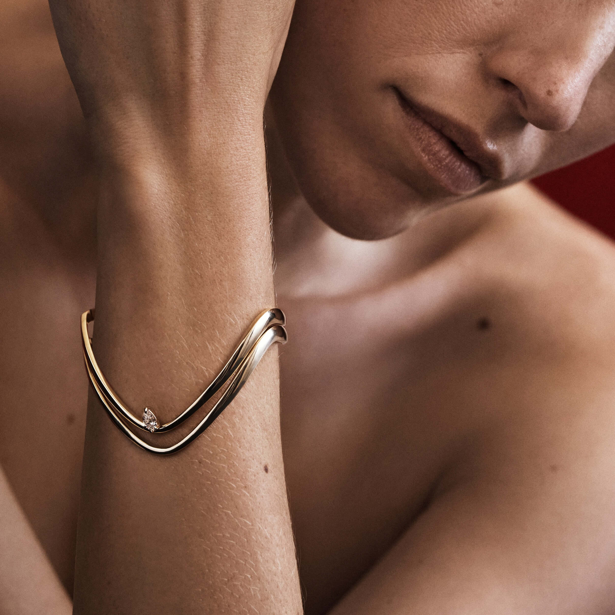 Gold bracelet on a wrist with a blurred background 
[text overlay: Model wears Piece 5—The Forme Bangle above Piece 8—The Sol Bangle]