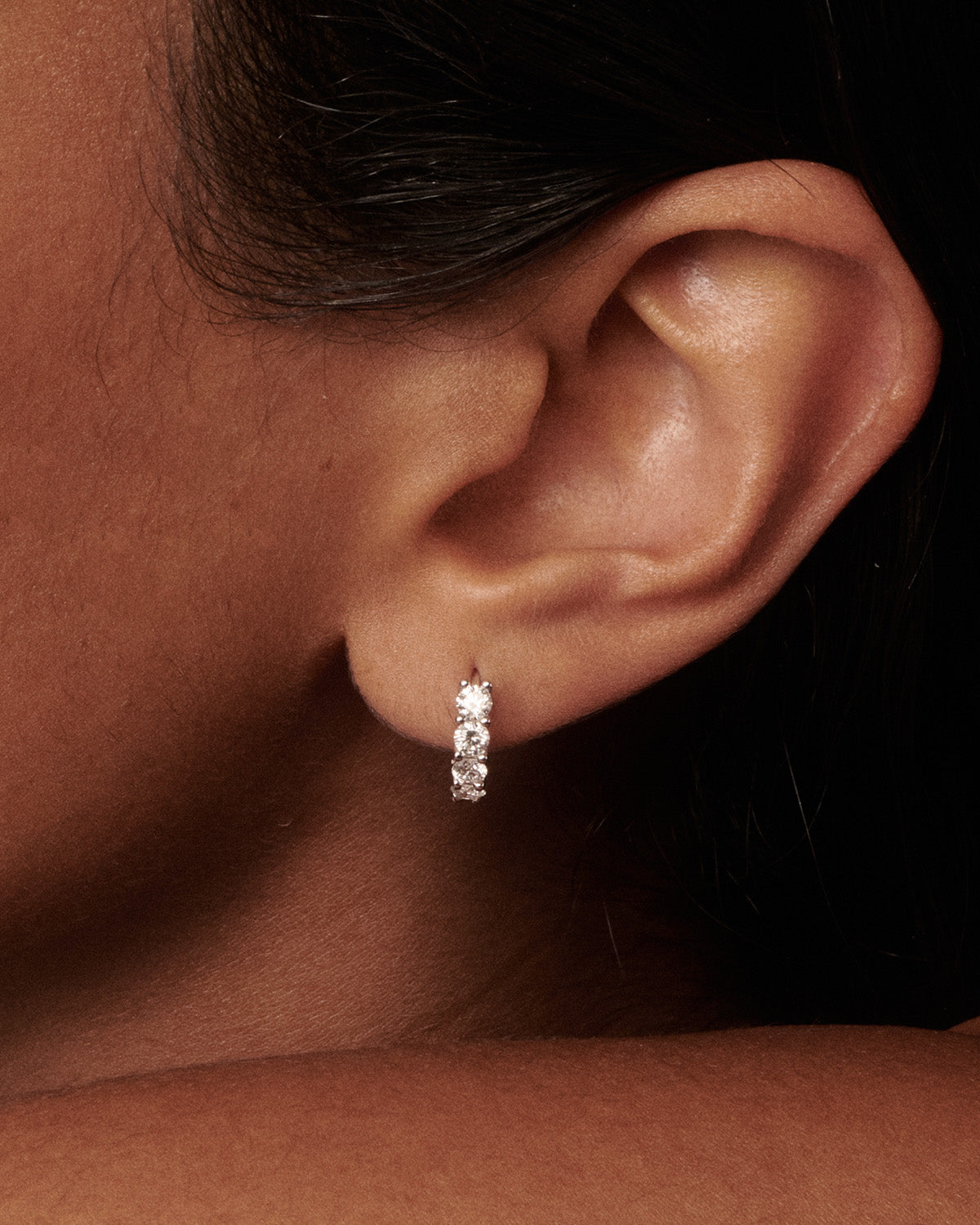 A close-up of a person's ear wearing a small silver hoop earring with three clear stones, set against dark hair and a dark background.