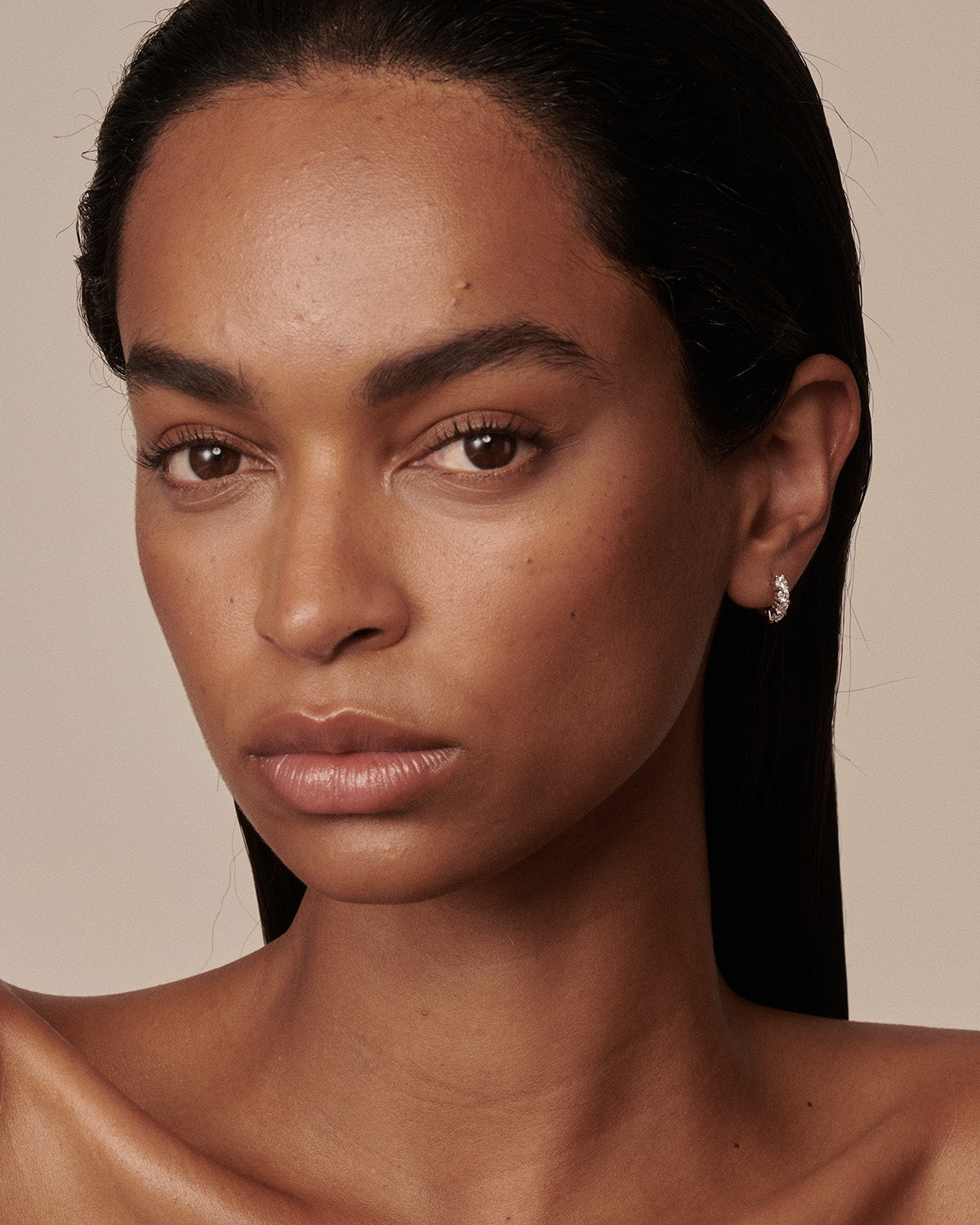 A close-up portrait of a model with dark hair pulled back, wearing a small silver crescent moon stud earring on her ear, against a solid beige background.