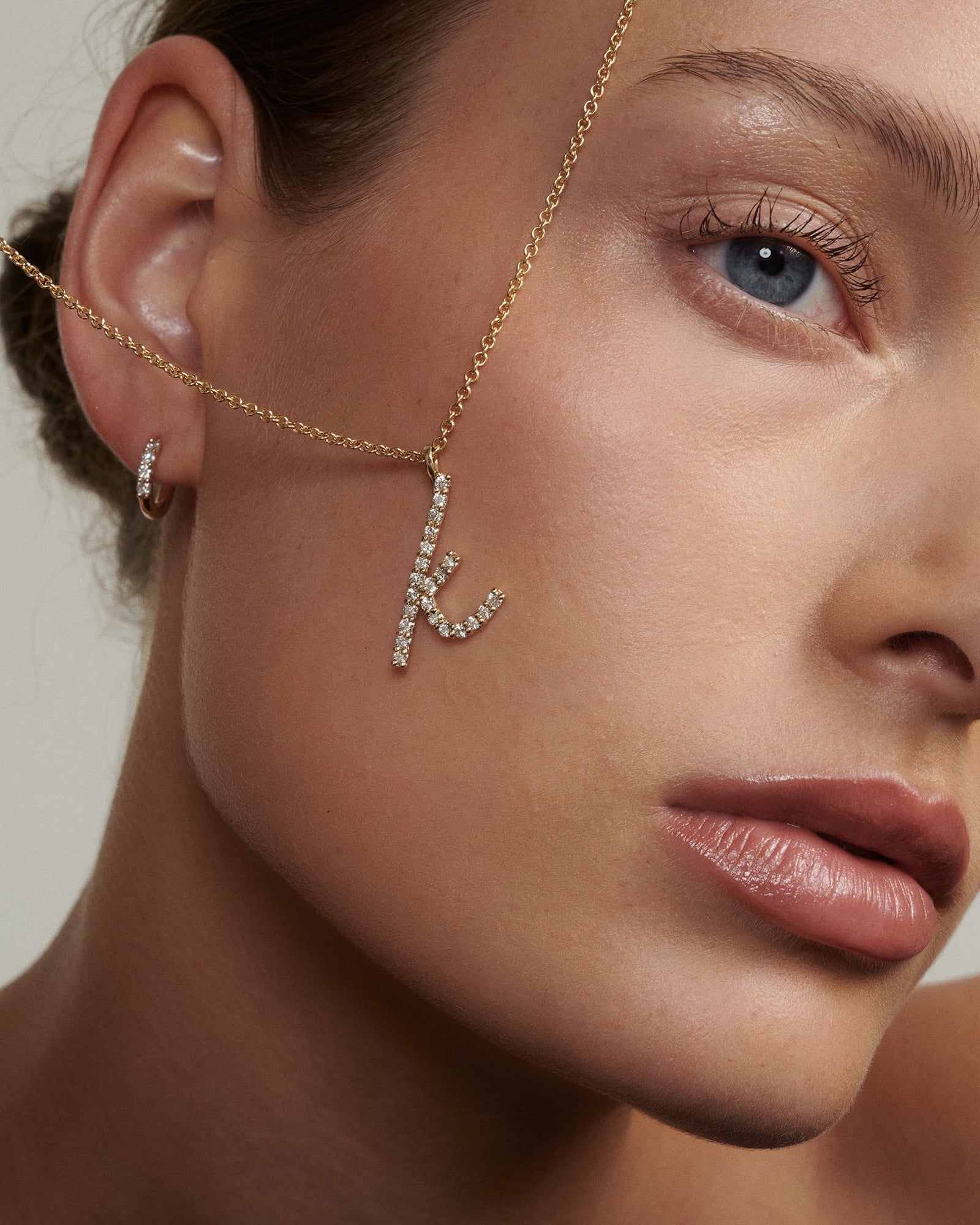 Close-up of a woman's face wearing a gold chain with a diamond letter 'K' pendant