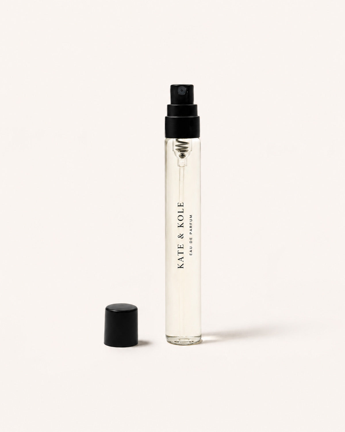 A clear glass perfume atomizer bottle with a black spray top and detached black cap, standing on a light warm off-white background. The bottle is labeled 'KATE & KOLE' and 'EAU DE PARFUM'.