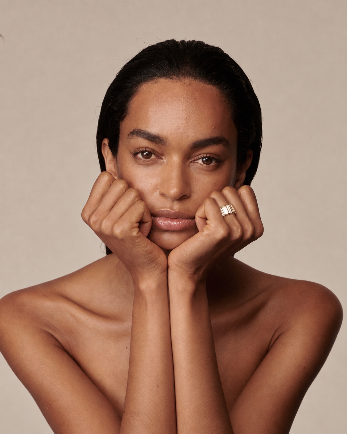 A close-up portrait of a woman with dark hair pulled back, resting her chin on her hands, wearing a wide gold band ring on one finger, set against a solid beige background.