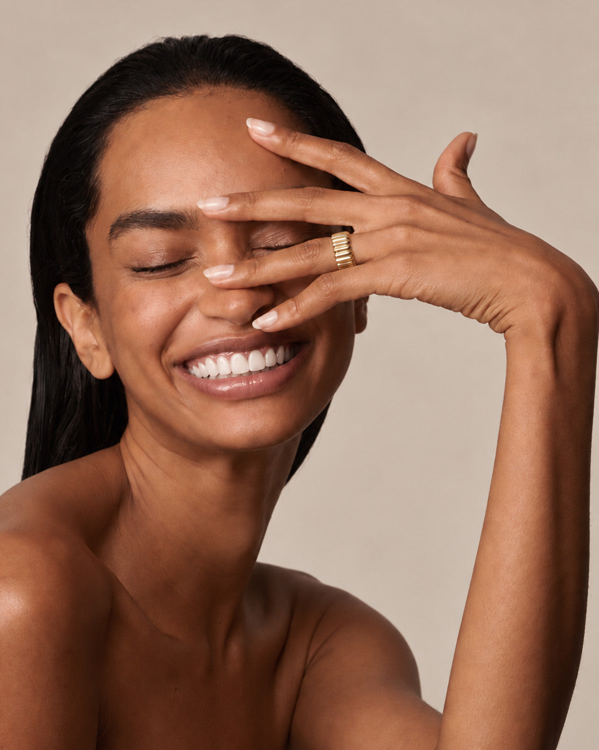 A close-up of a smiling woman with her hand covering part of her face, wearing a gold ring with a textured, ridged band on her finger, set against a solid beige background.