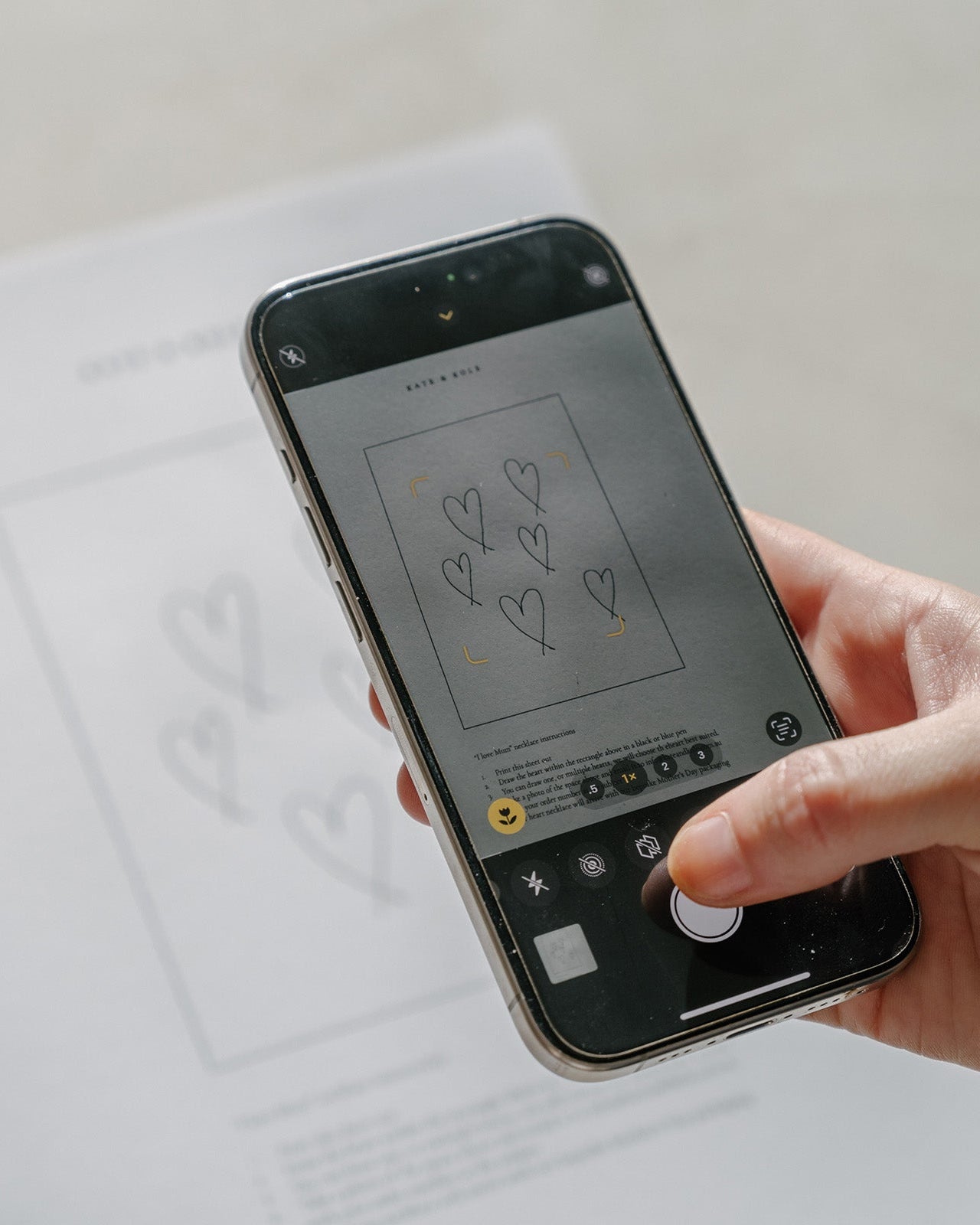 Hand holding a smartphone, capturing a photo of a paper with heart designs, possibly related to luxury jewellery sketches.