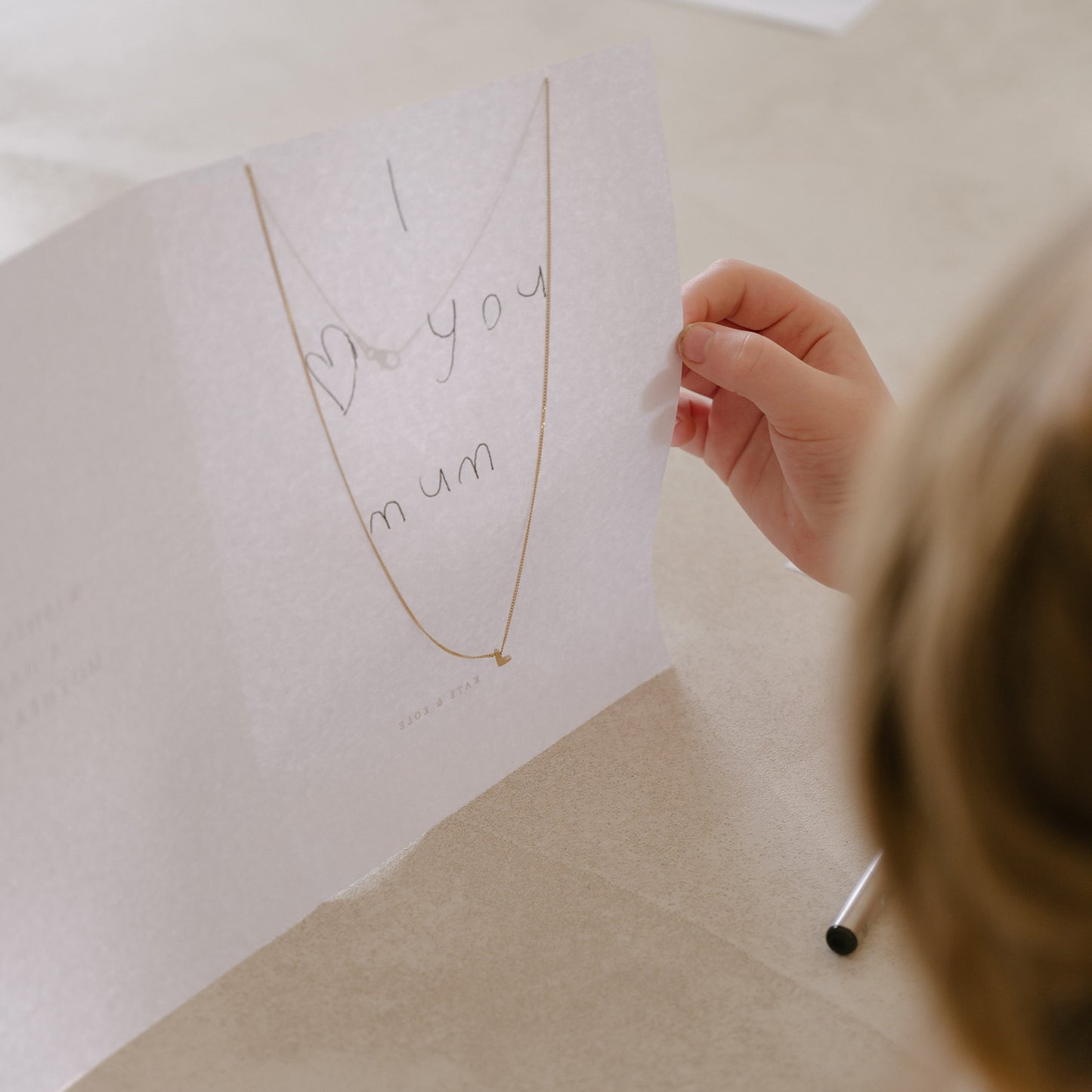 Child's hand holding a card with "I love you mum" and a delicate gold necklace, symbolising luxury and affection in a heartfelt gift.