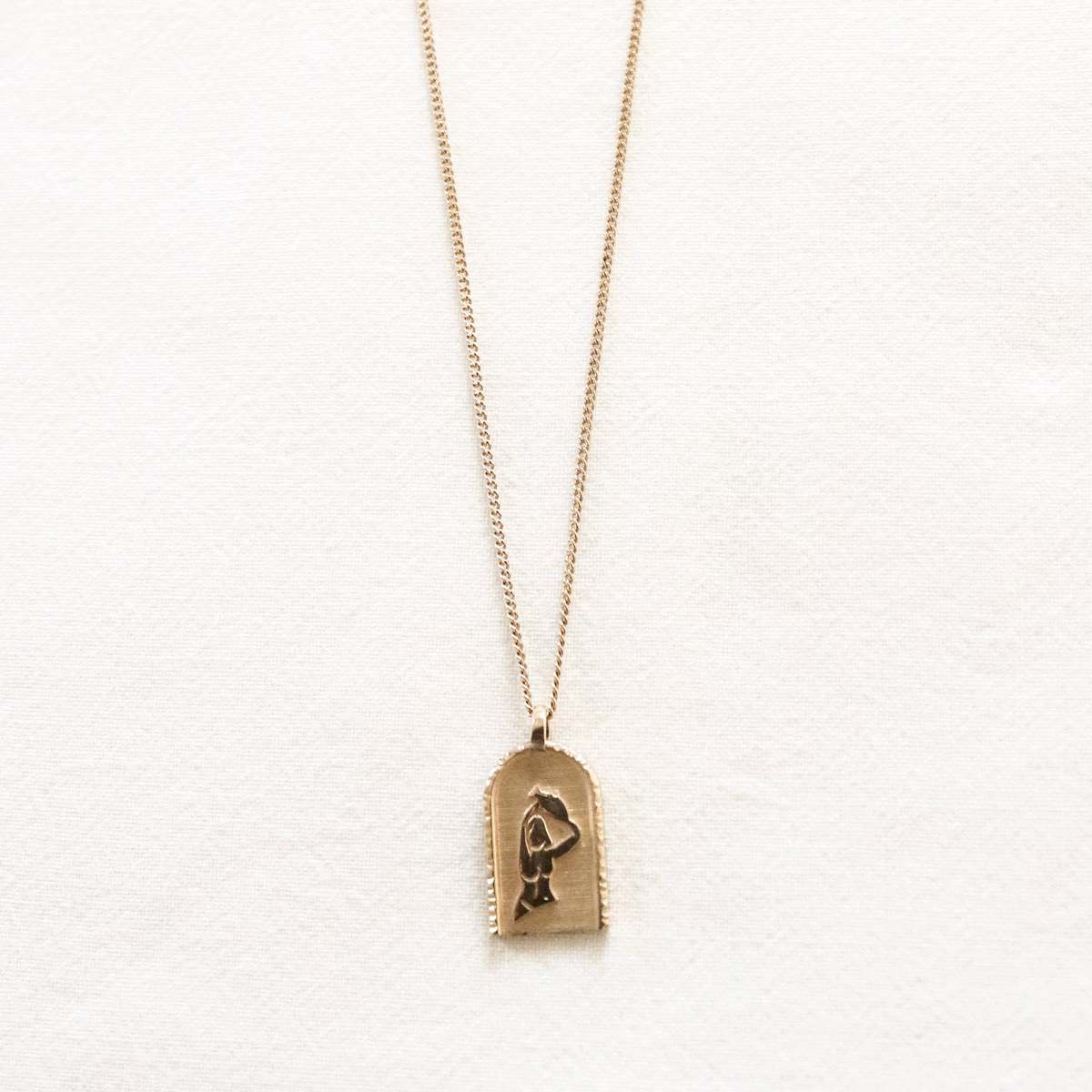 Gold necklace with rectangular pendant featuring an engraved figure. Set on a light background, showcasing luxury jewellery.