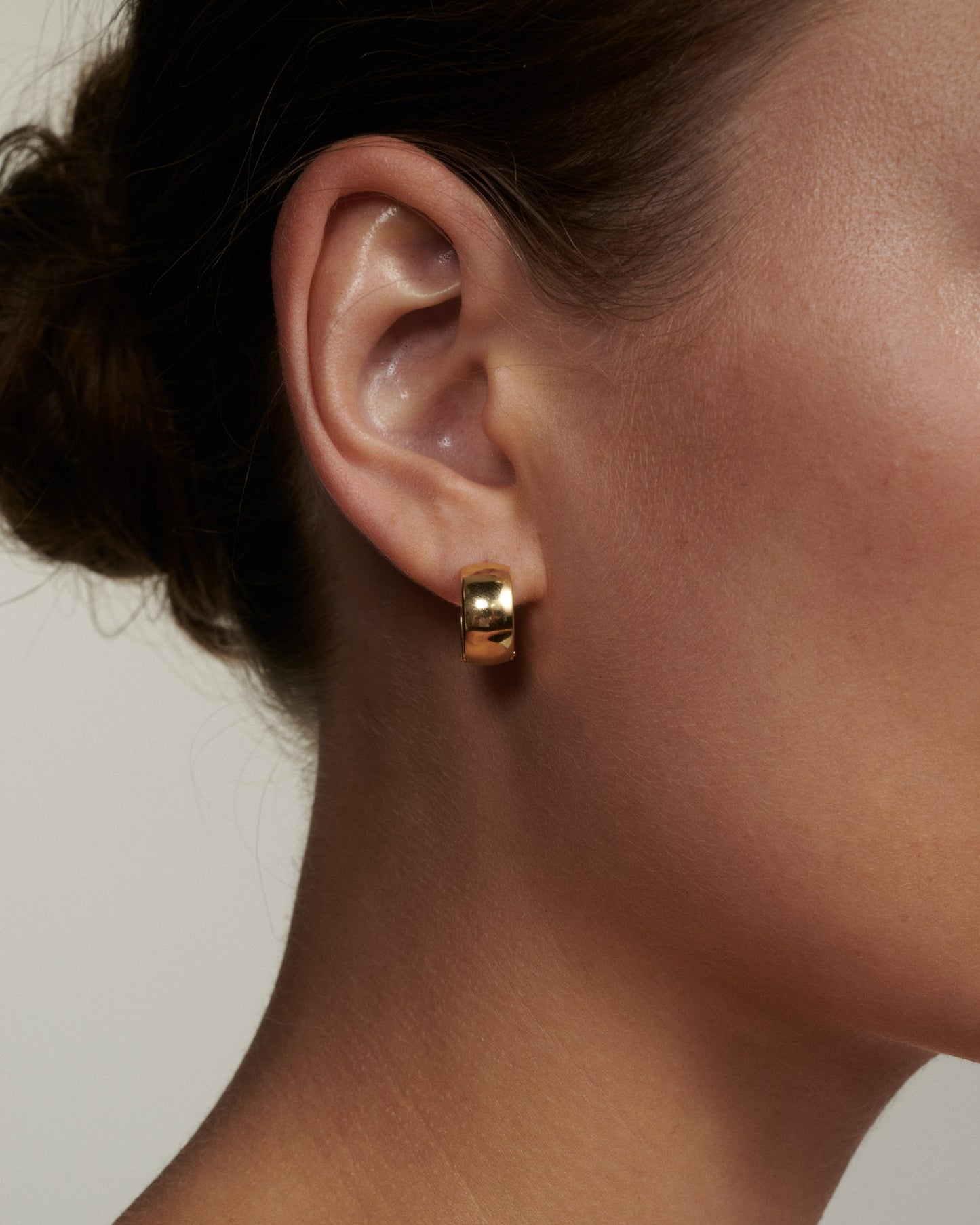 Close-up of an ear wearing a gold hoop earring against a neutral background