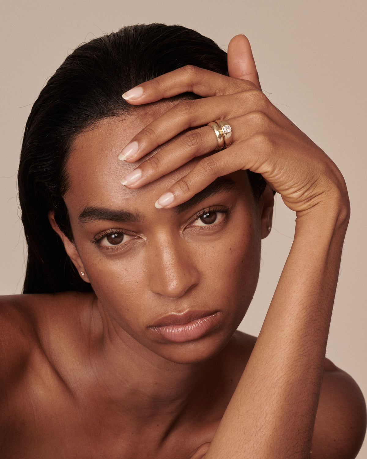 A close-up portrait of a woman with dark hair, wearing a gold ring with a cushion cut central diamond on her left hand, posed against a solid beige background.