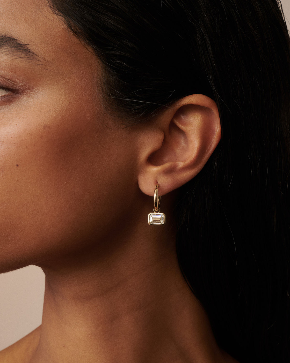 A close-up of a model's ear wearing a delicate gold huggie hoop earring with a small rectangular clear gemstone  dangle. [text overlay: Francielle wears 4ct total carat weight]