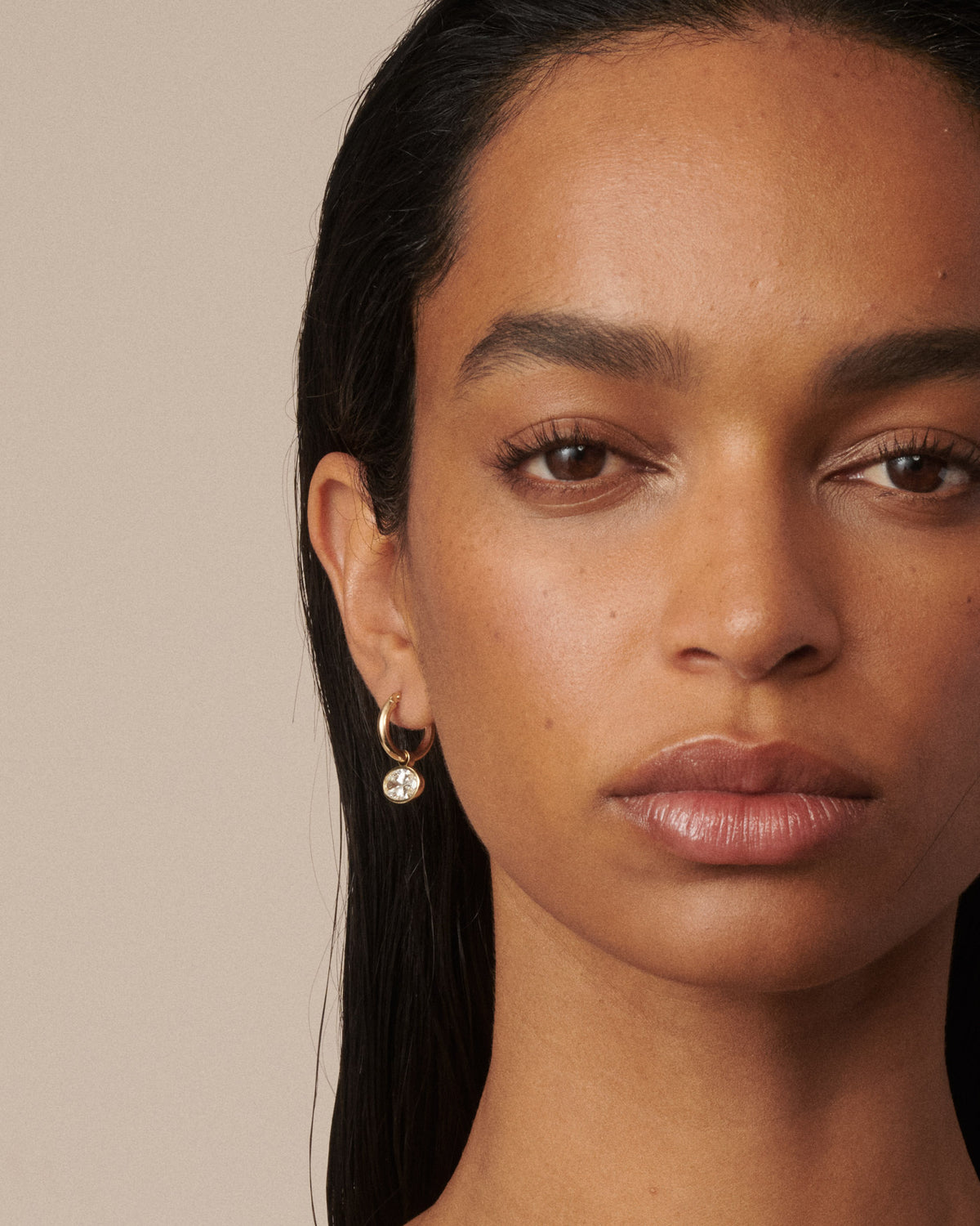 A close-up portrait of a model wearing a delicate gold hoop earring with a small clear diamond pendant against a solid beige background.