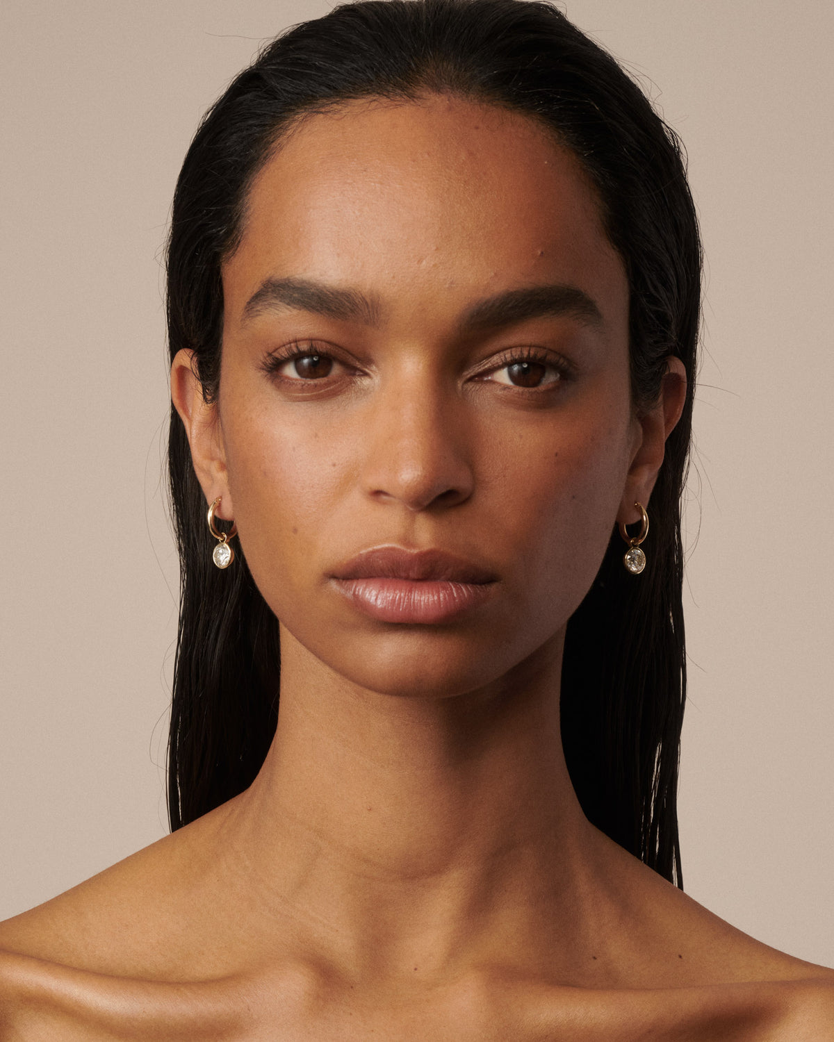 A close-up portrait of a woman with dark hair wearing small gold hoop earrings with clear diamond drops, set against a solid beige background. [text overlay: Francielle wears 4ct total carat weight]