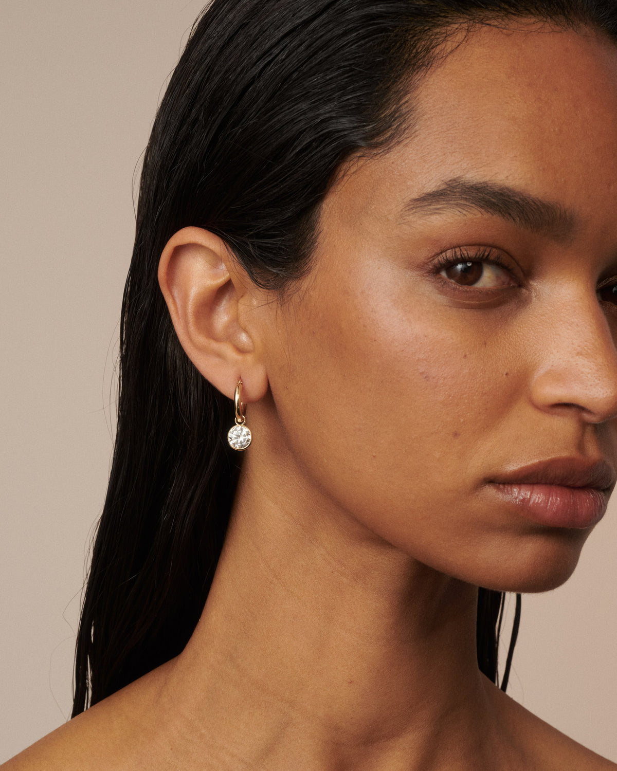 A close-up of a model wearing a delicate gold hoop earring with a small clear faceted gemstone dangle against a warm beige background.