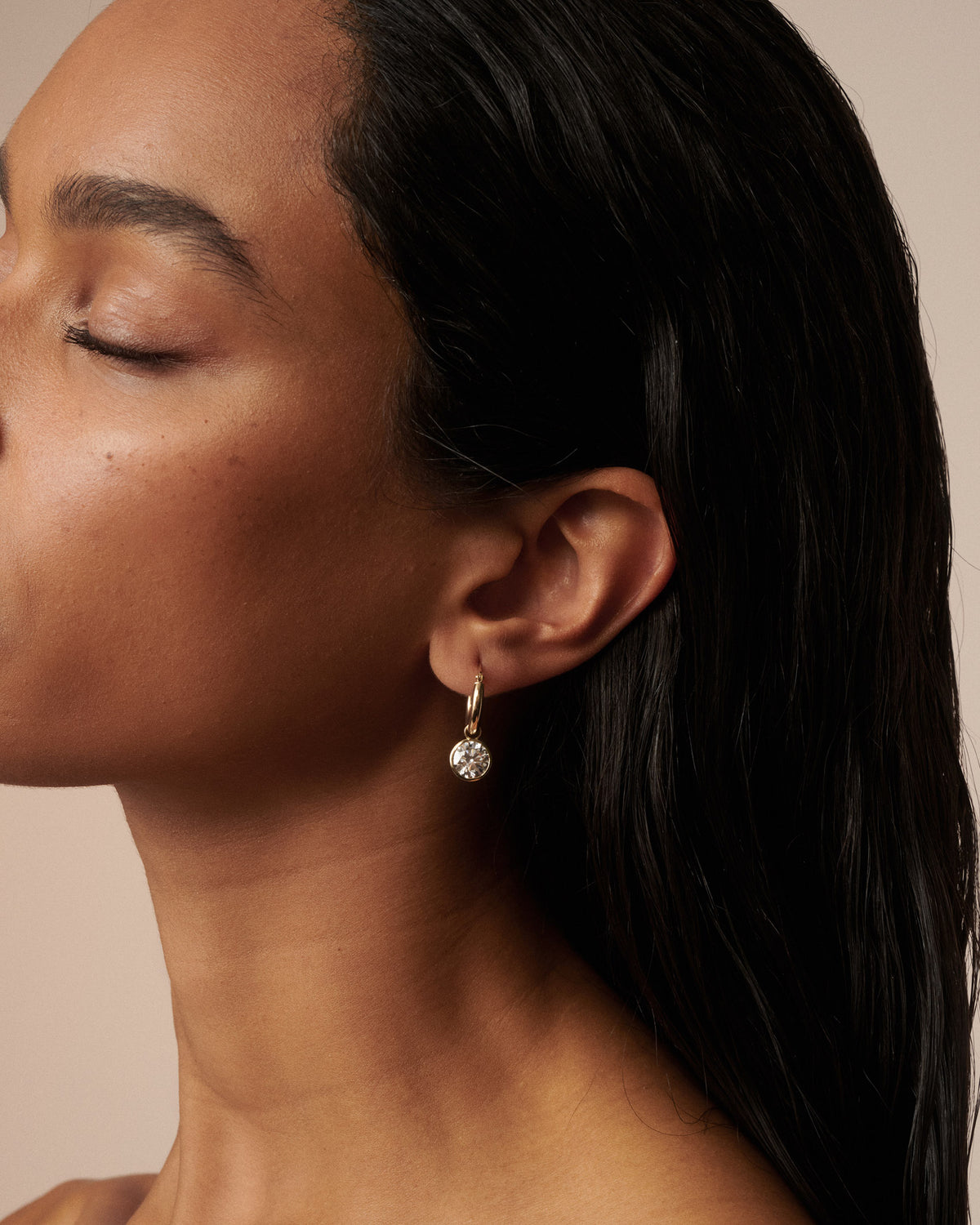 A close-up side profile of a woman with closed eyes wearing a delicate gold hoop earring with a small clear faceted drop stone, set against a warm beige background. [text overlay: Francielle wears 4ct total carat weight]
