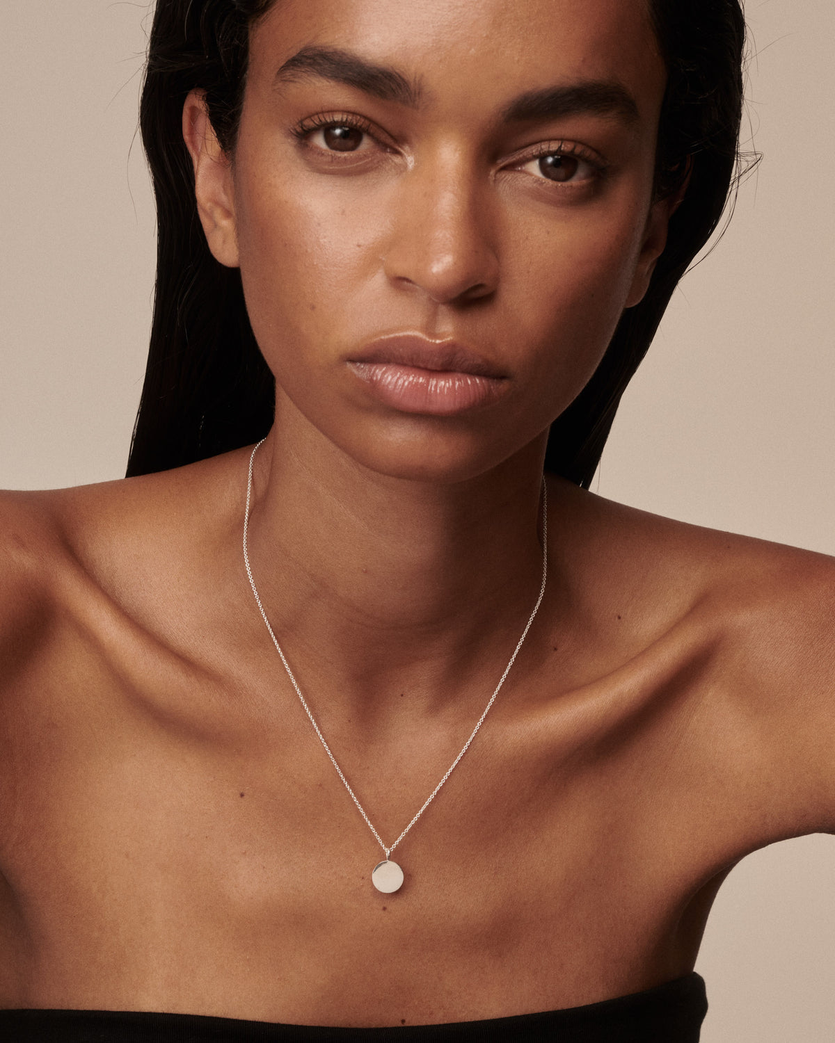 A close-up of a model wearing a delicate silver chain necklace with a small, round, disc-shaped pendant on a warm beige background. [text overlay:Large Pendant]
