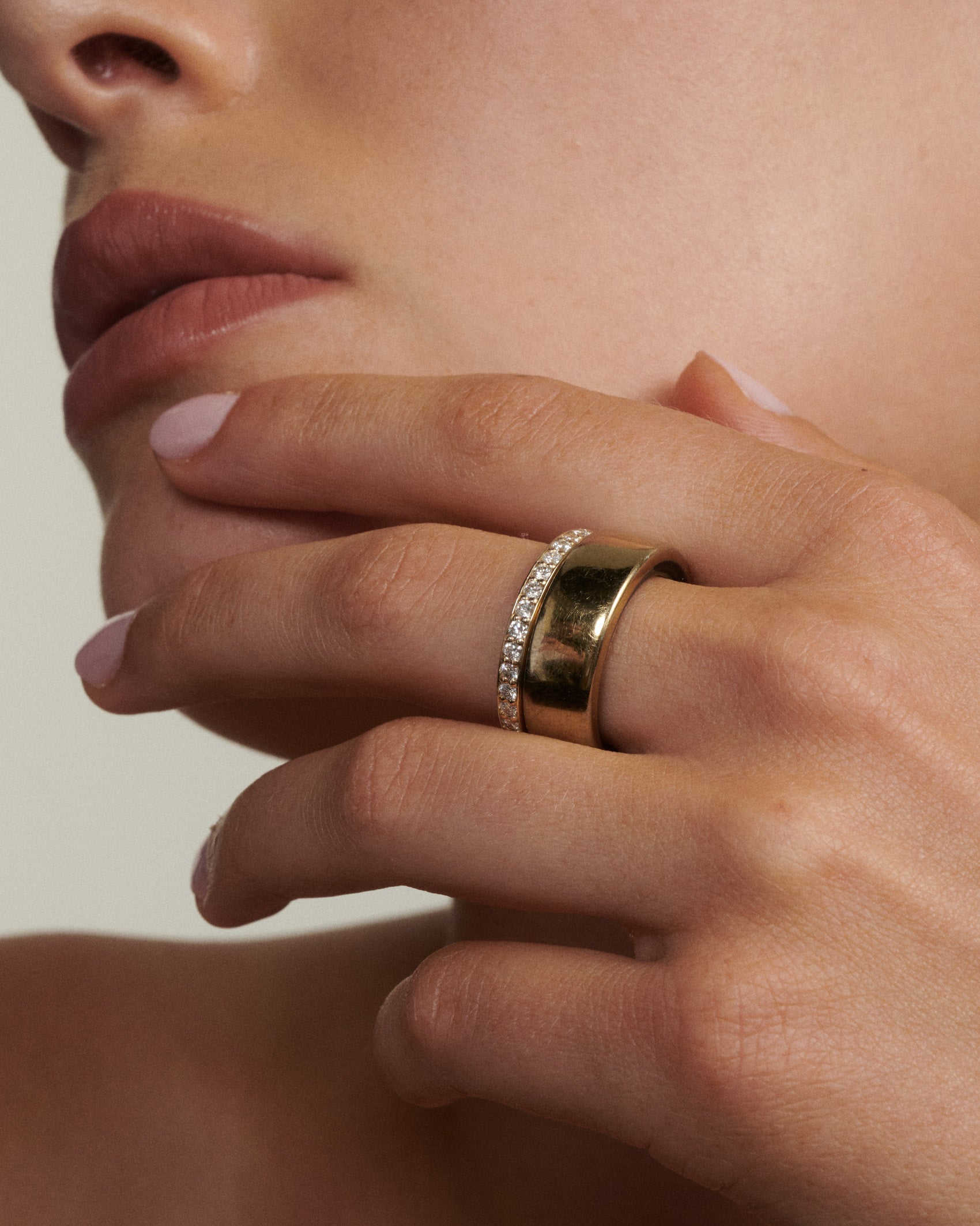 Close-up of a hand wearing two gold rings with a neutral background  [text overlay: Andrea wears the Half Set Wellness Ring (9ct Yellow Gold, 7) stacked with the Oura Ring]