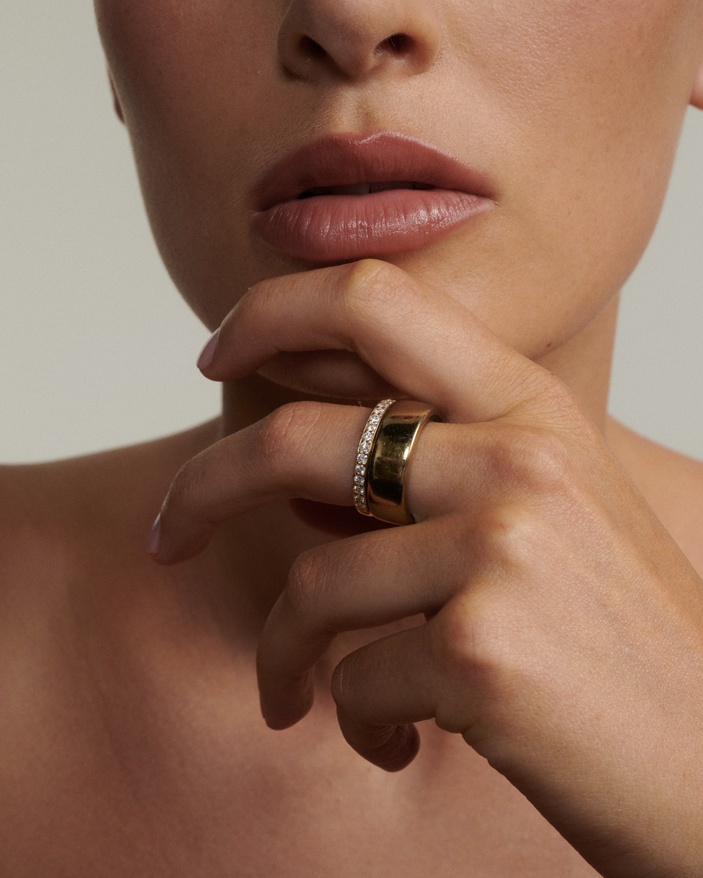 Close-up of a person wearing a gold ring on their finger with a neutral background  [text overlay: Andrea wears the Half Set Wellness Ring (9ct Yellow Gold, 7) stacked with the Oura Ring]
