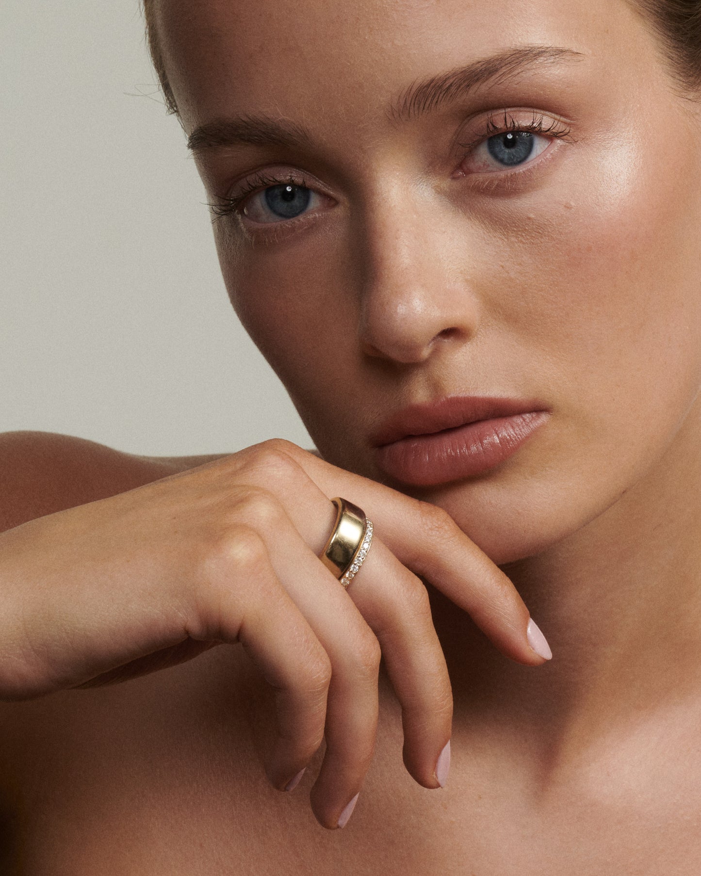 Close-up of a woman's face with a gold ring on her finger against a neutral background  [text overlay: Andrea wears the Half Set Wellness Ring (9ct Yellow Gold, 7) stacked with the Oura Ring]