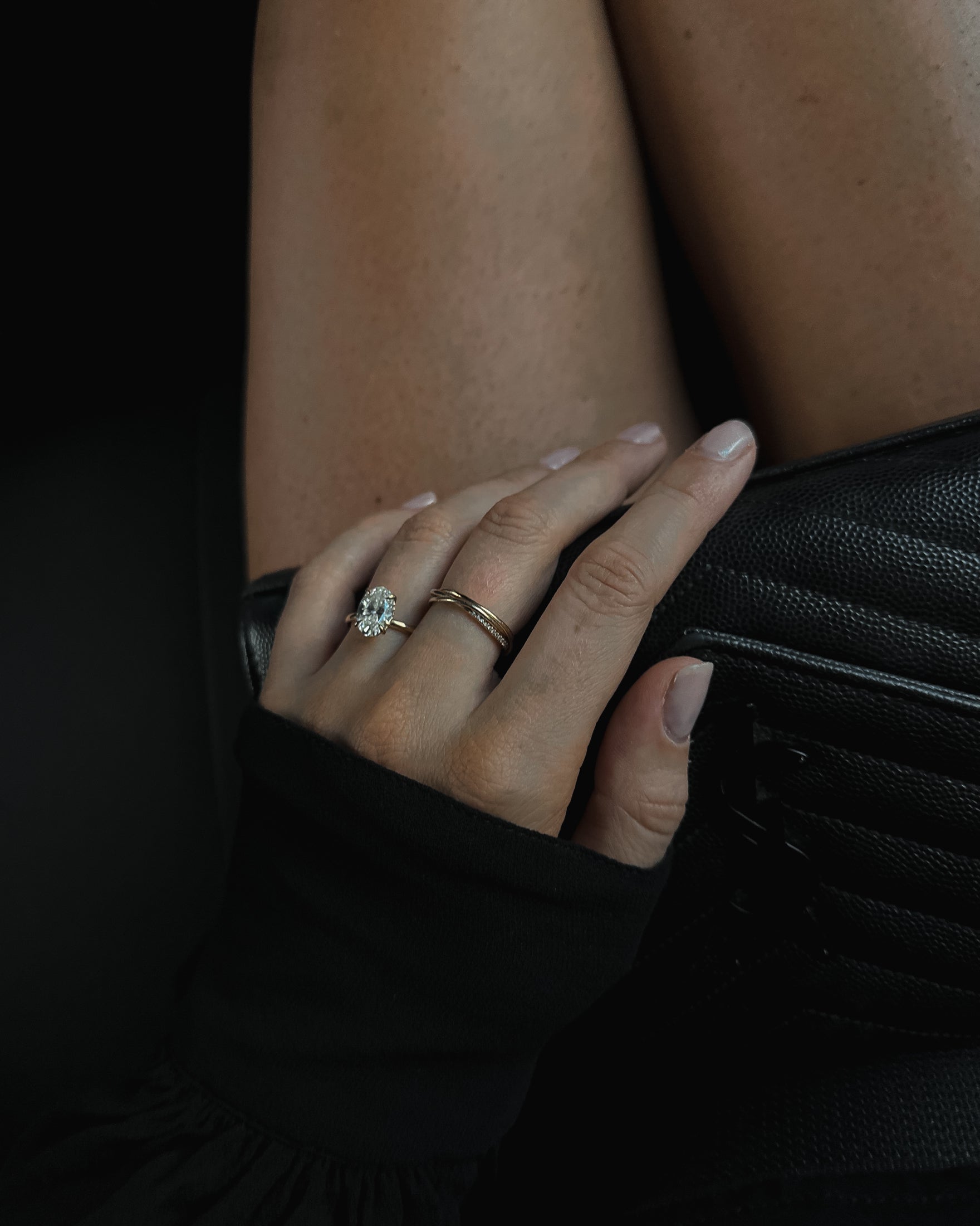 A hand with a diamond ring and gold band rests on a black leather handbag, with bare legs in the background. Luxurious jewellery showcase.