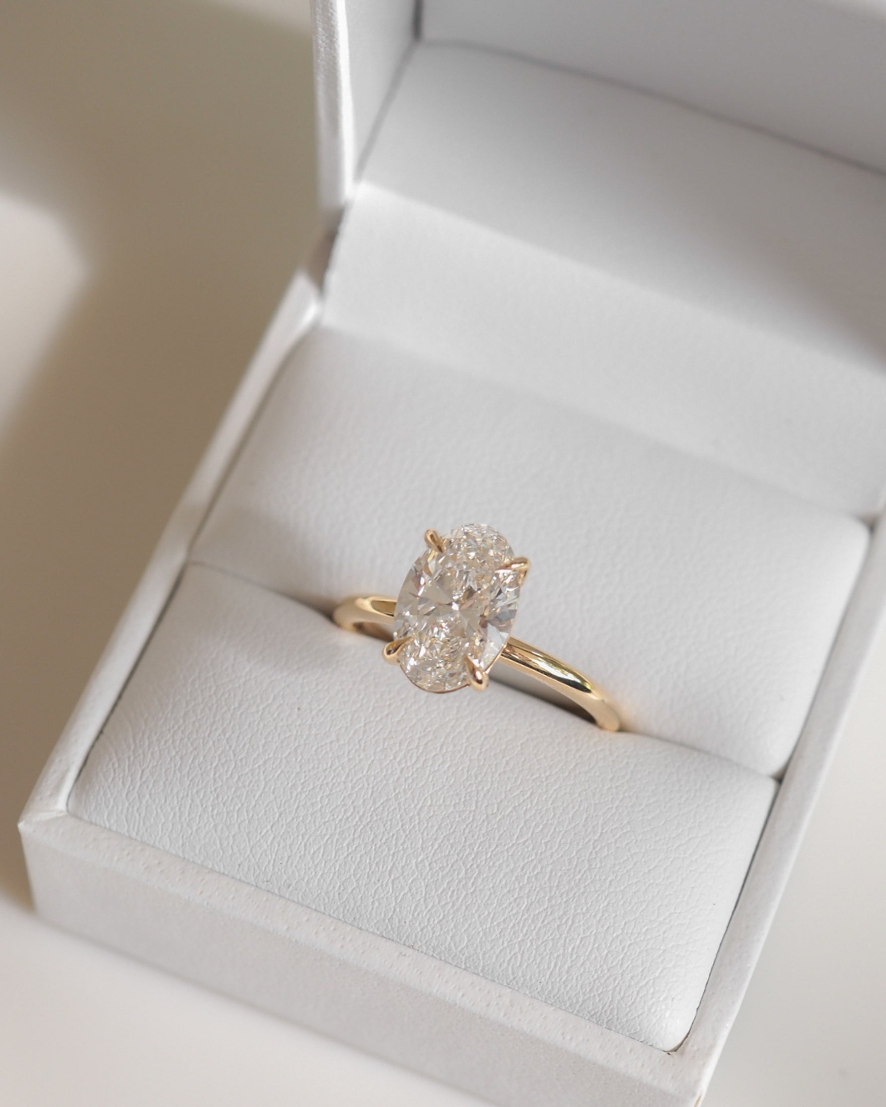 Oval diamond solitaire ring in a simple gold band, displayed in an open white jewellery box, on a light background.