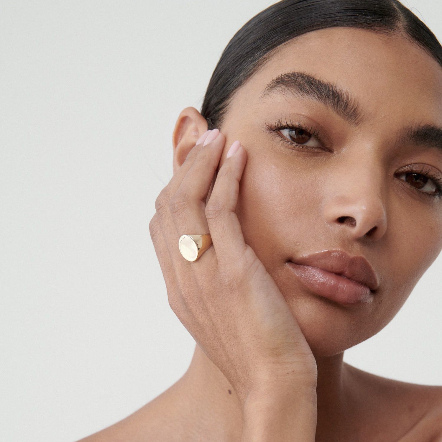 Woman with sleek hair wearing a gold statement ring, hand on face, showcasing luxury jewellery against a plain background.