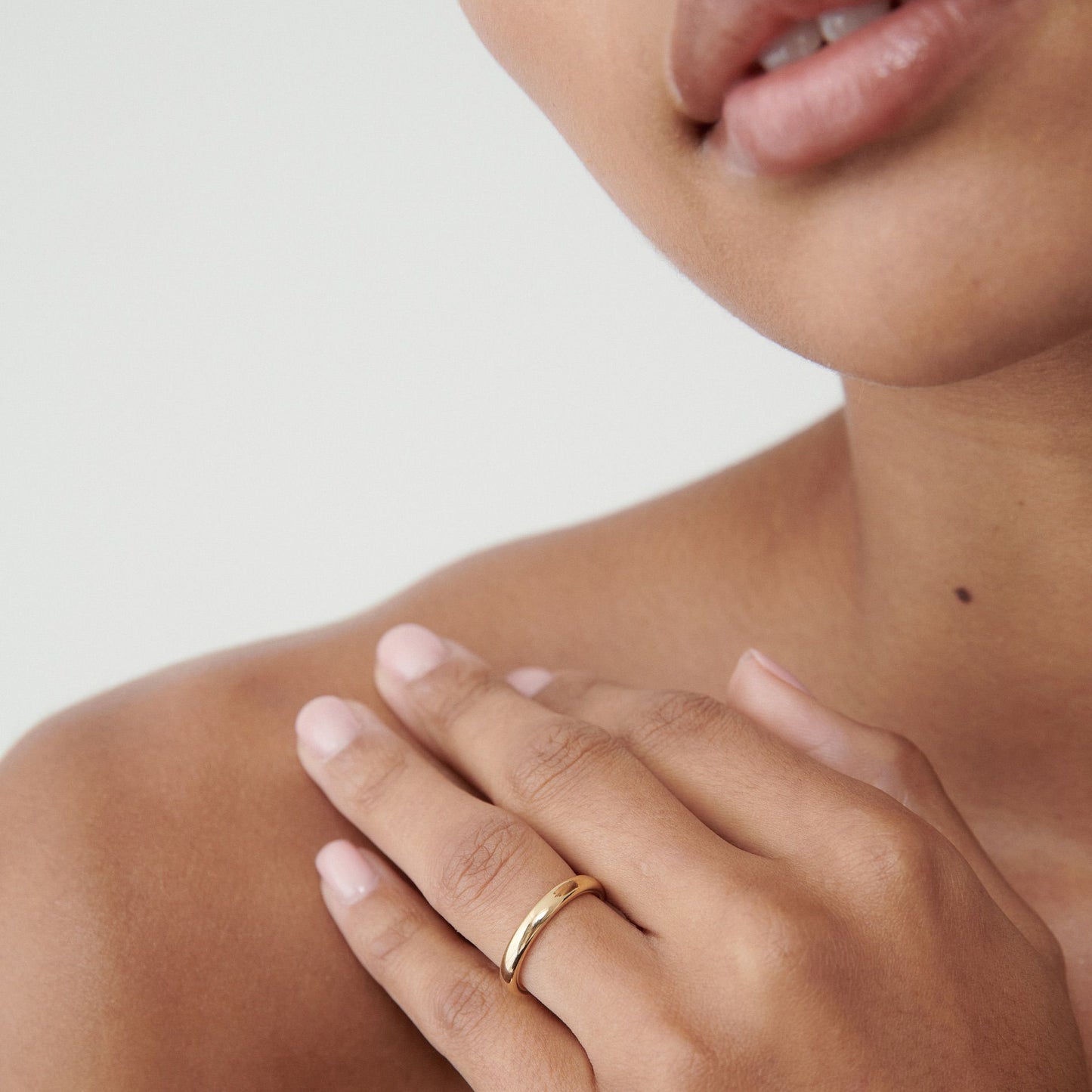 Close-up of a woman touching her shoulder, wearing a sleek gold band on her finger. Her lips are slightly parted, conveying elegance.
