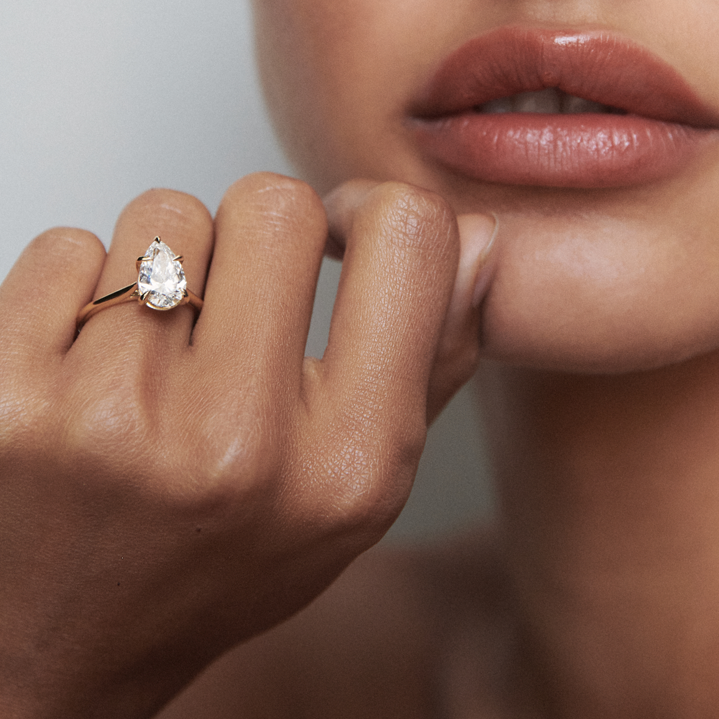 Close-up of a woman's hand with a pear-shaped diamond ring on a gold band near her lips. Elegant luxury jewellery style.
