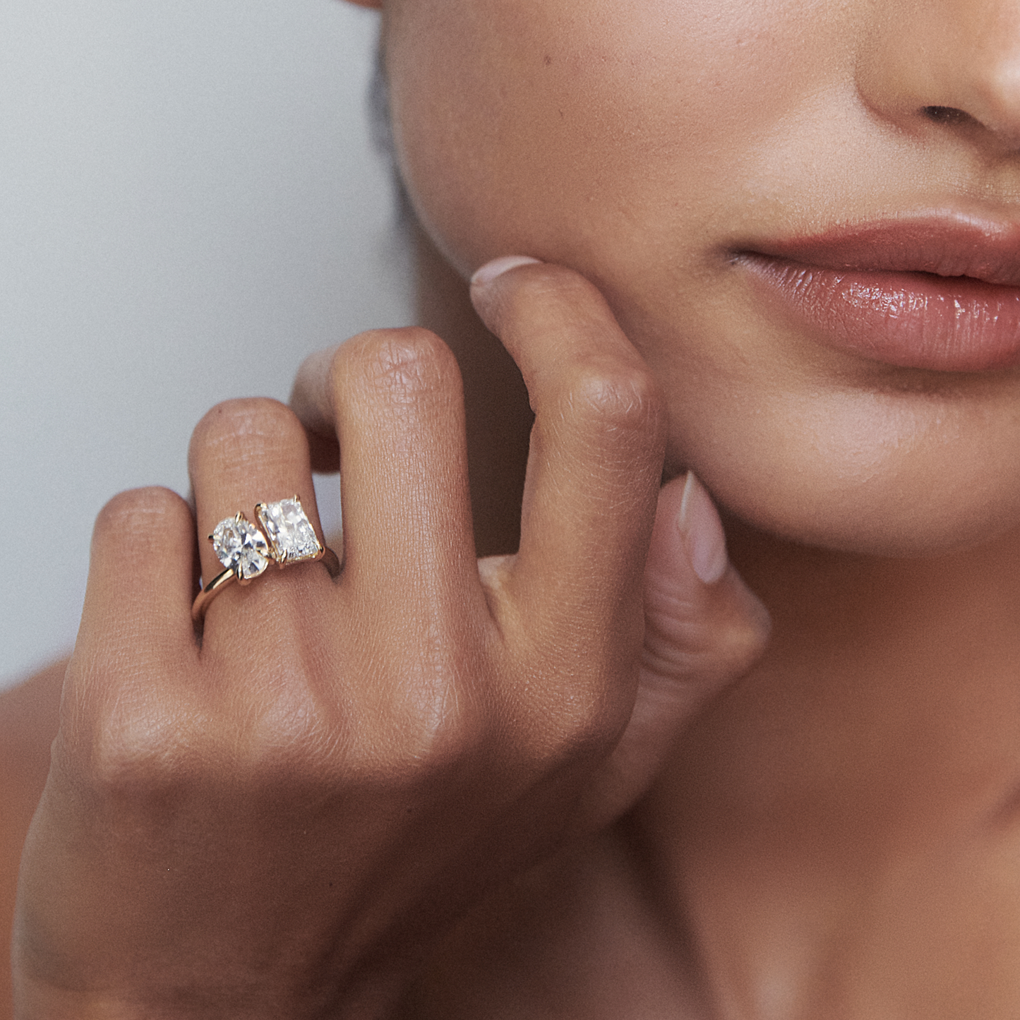 Close-up of a hand wearing a luxury gold ring with an oval and square diamond, gently resting on a woman's face, showcasing elegant jewellery design.