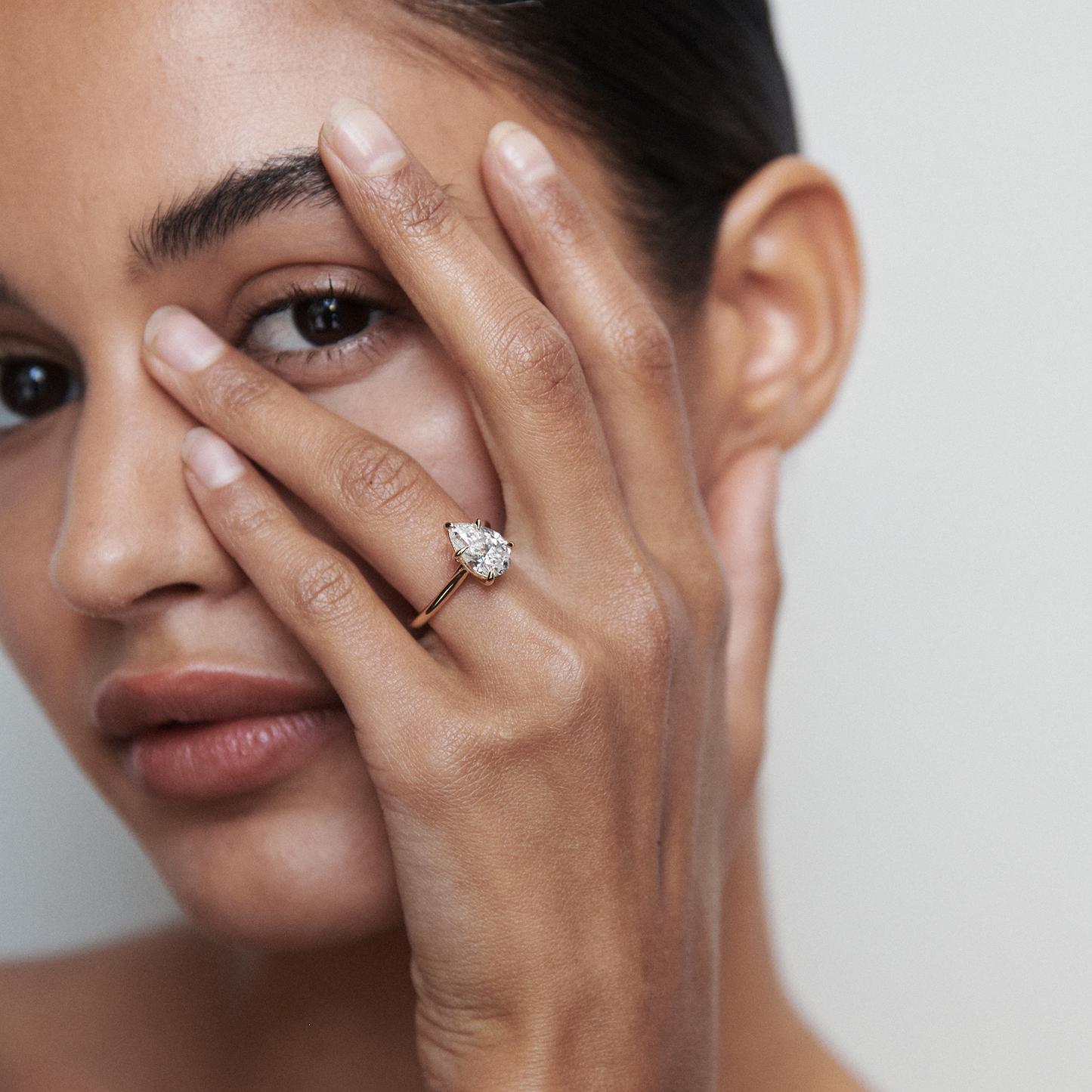 Woman with sleek bun poses, displaying a pear-shaped diamond ring on her finger, close to her face. Luxurious jewellery detail.