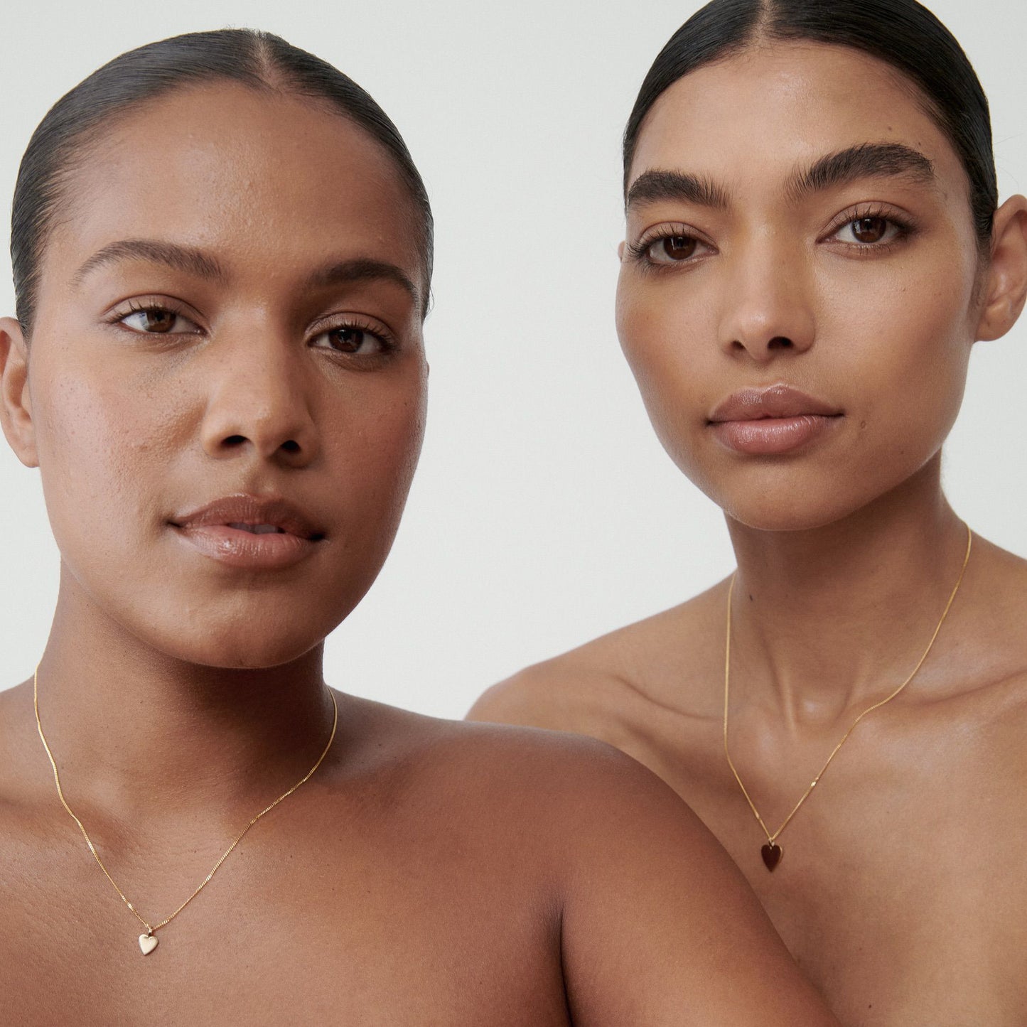 Two women wearing elegant gold necklaces with heart pendants; bare shoulders and neutral backgrounds emphasise luxury jewellery design.