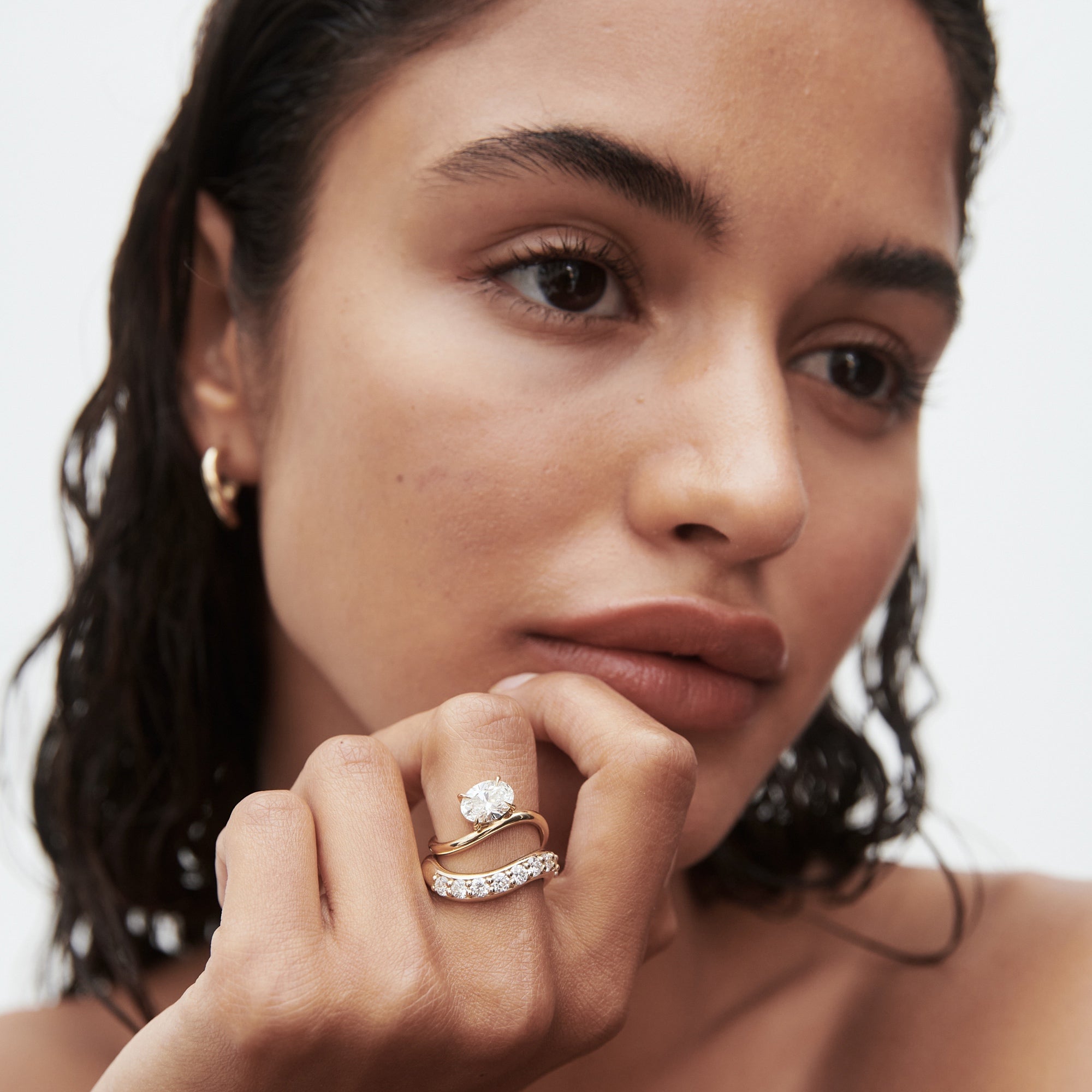 Woman wearing elegant gold and diamond rings, posing thoughtfully with hand near face. Luxury jewellery details stand out beautifully.