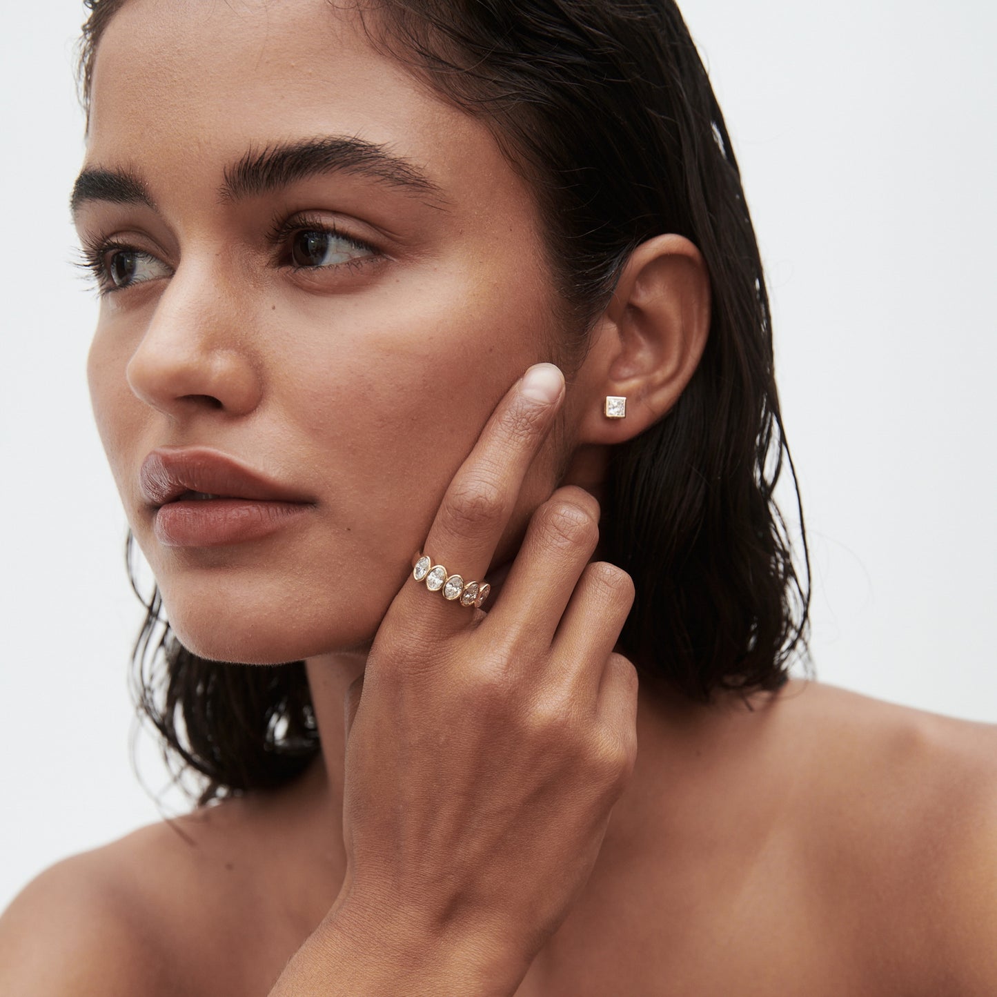 Woman with wet hair wears a gold diamond stud earring and a gold ring with clustered gemstones, set against a plain background.