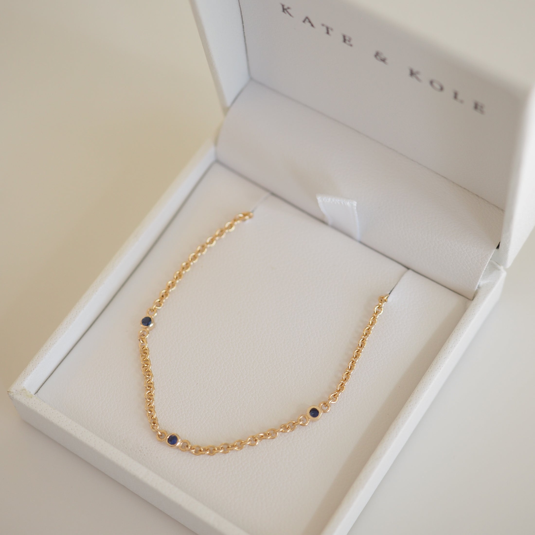Gold bracelet with blue gemstones in a white Kate & Kole jewellery box. Elegant luxury accessory, perfect for sophisticated style.