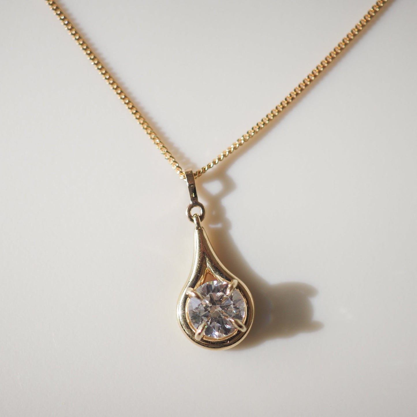 Gold necklace featuring a teardrop pendant with a sparkling diamond, set on a fine chain, casting a soft shadow on a light background.