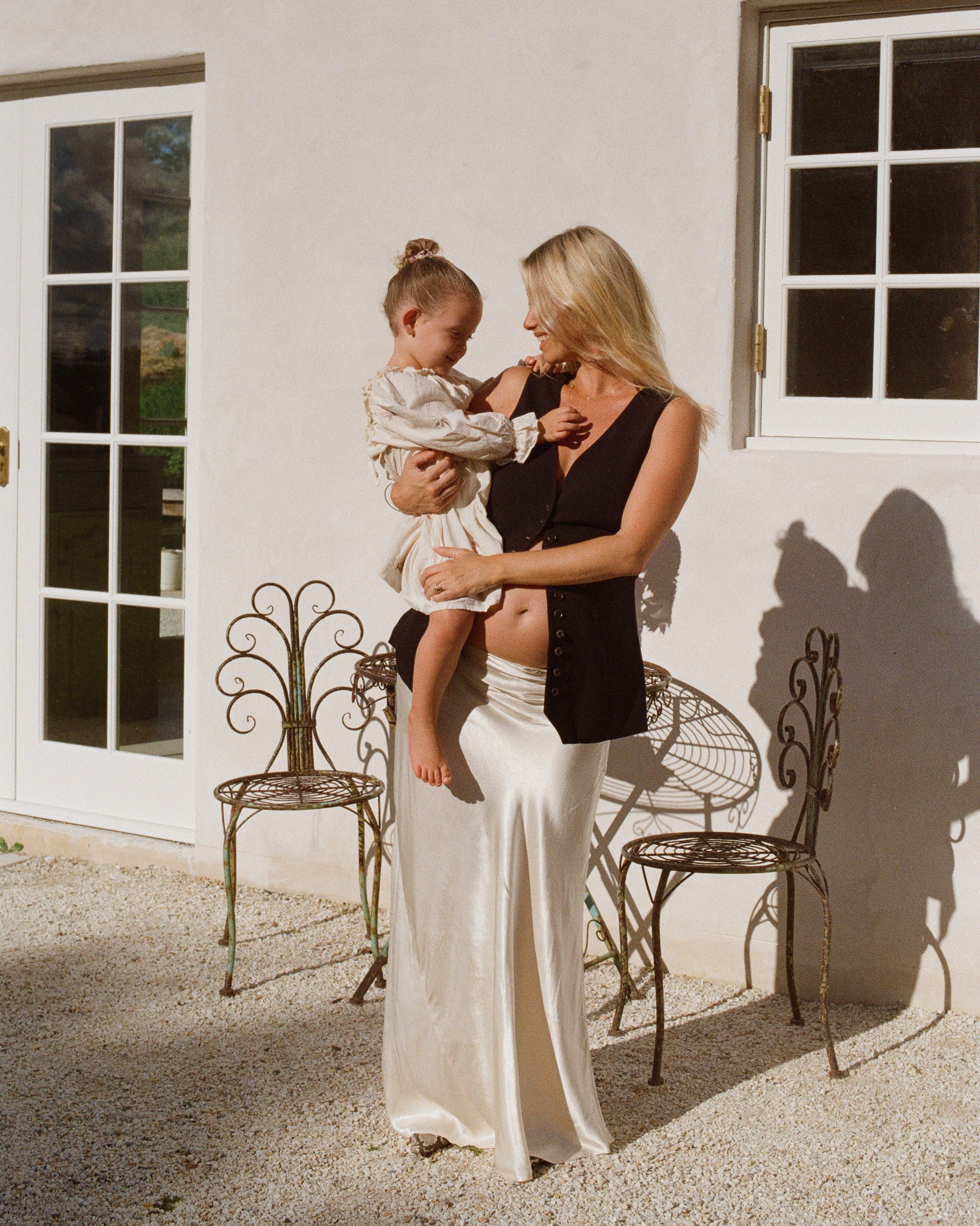 A woman in a sleeveless top and silk skirt holds a child outdoors by a white wall with decorative chairs. Sunlight creates gentle shadows.