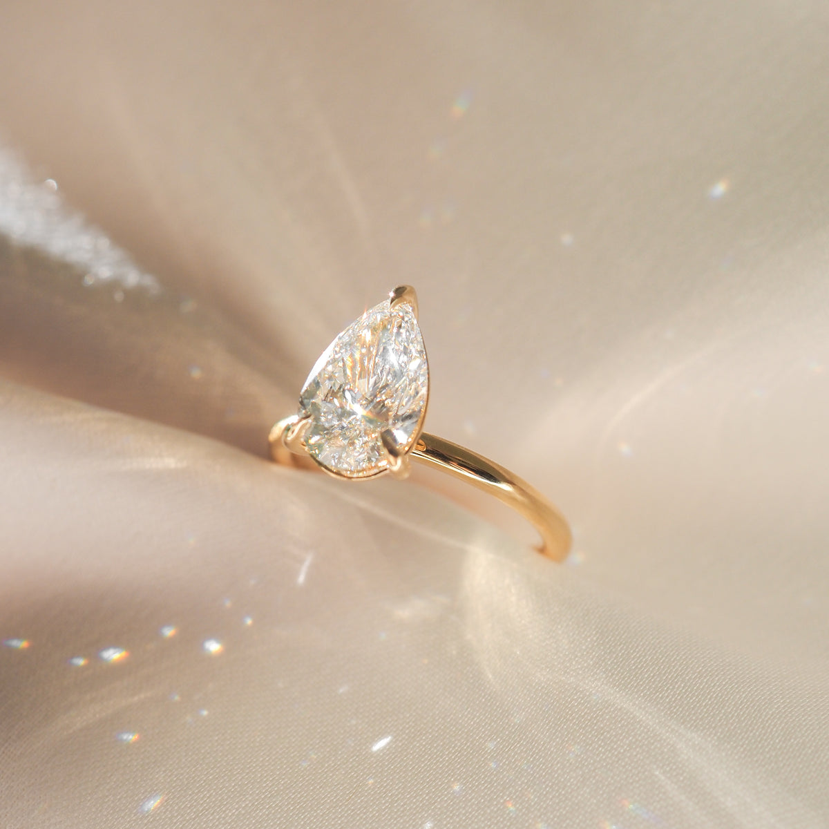 Pear-shaped diamond ring set in gold, elegantly displayed on a shimmering fabric backdrop.
