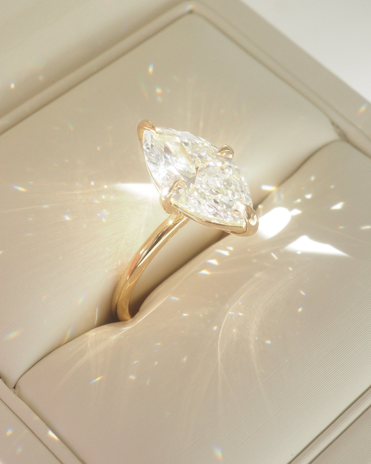 A yellow gold ring with a marquise-cut clear gemstone displayed in a cream-colored jewelry box.