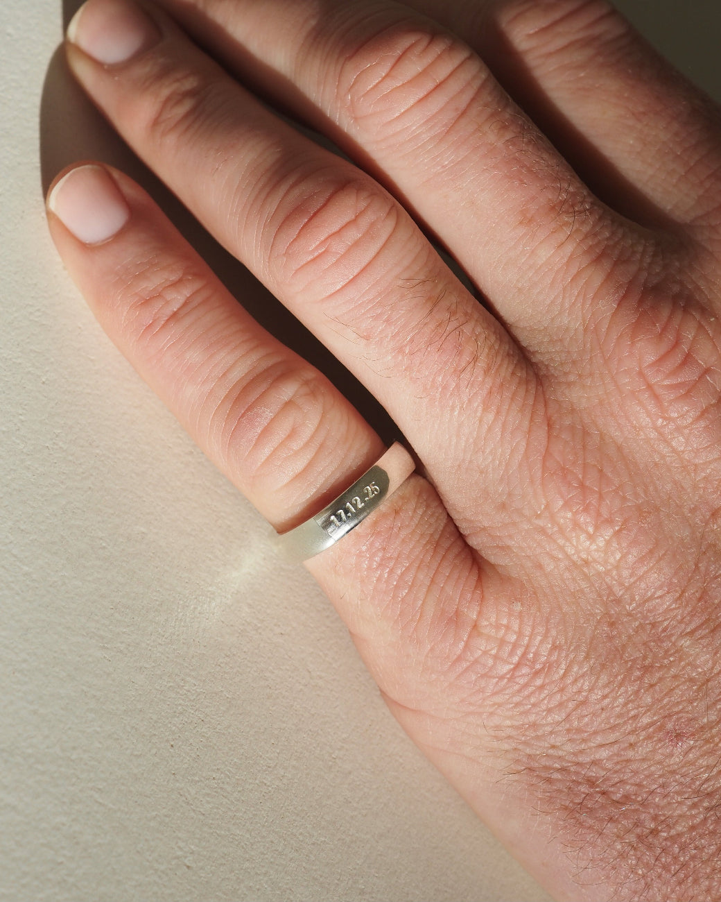 A close-up of a person's hand wearing a thin silver band ring engraved on the pinky finger, resting against a neutral beige background.