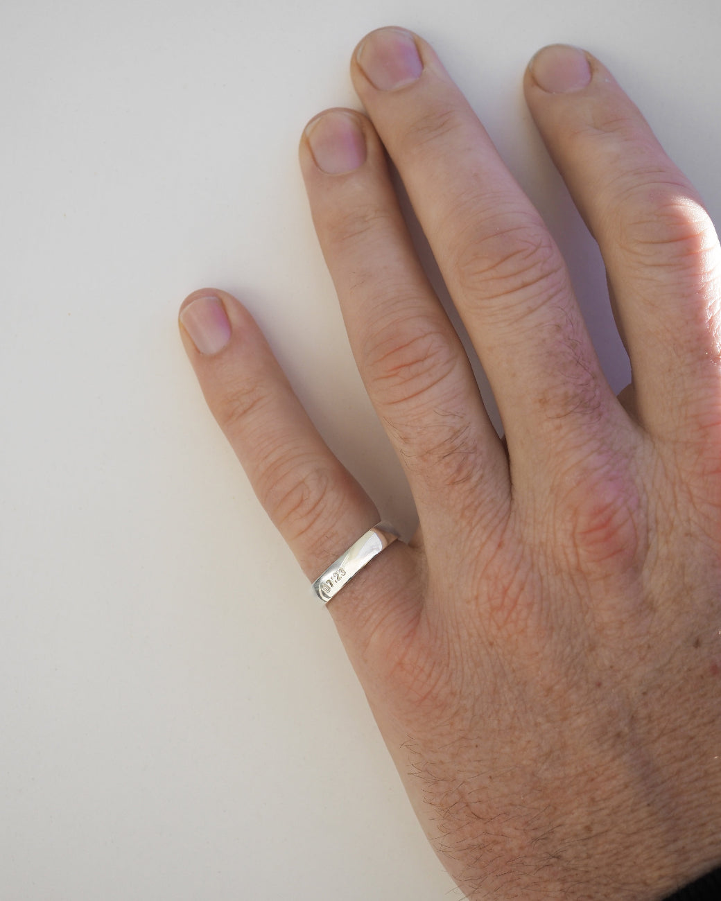 A close-up of a person's hand wearing a simple silver band ring on the ring finger, photographed against a plain light background.