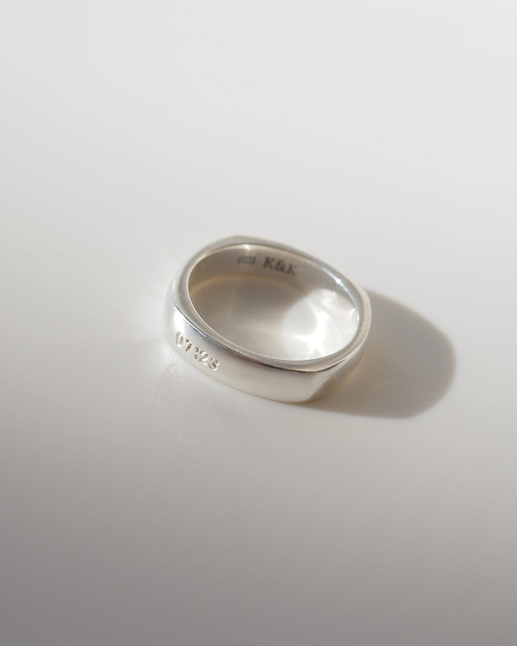 A polished sterling silver band ring with engraved markings, including 'K&K' on the inside and a date on the outside, resting on a light gray surface with a soft shadow.